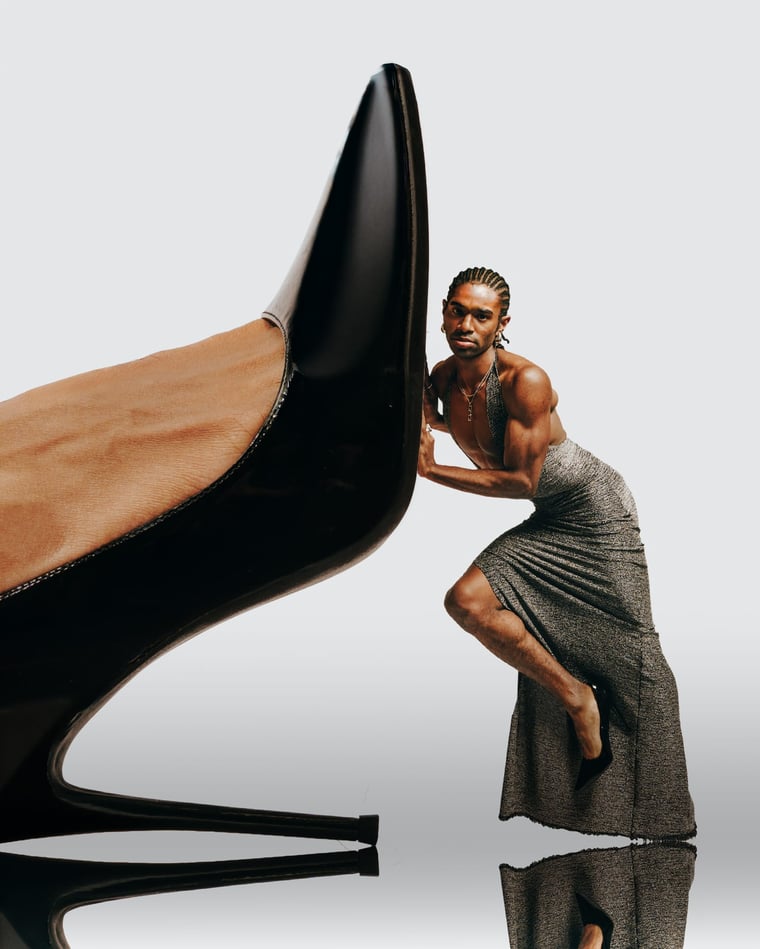 A dancer in a grey draped dress braced against an enormous black stiletto on a white sweep, dwarfed by it
