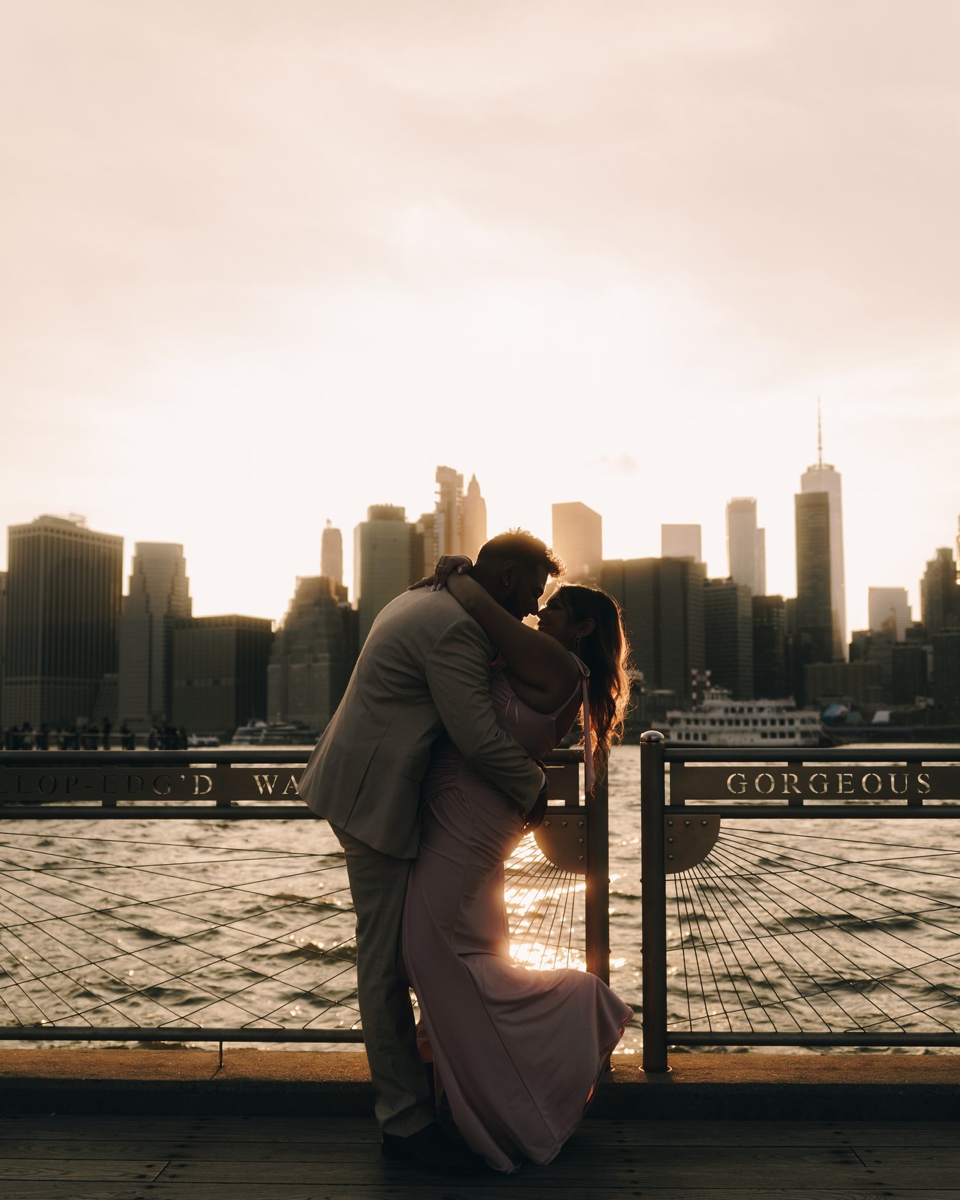 A couple kissing against the railing of a waterfront promenade, the towers of lower Manhattan going gold behind them