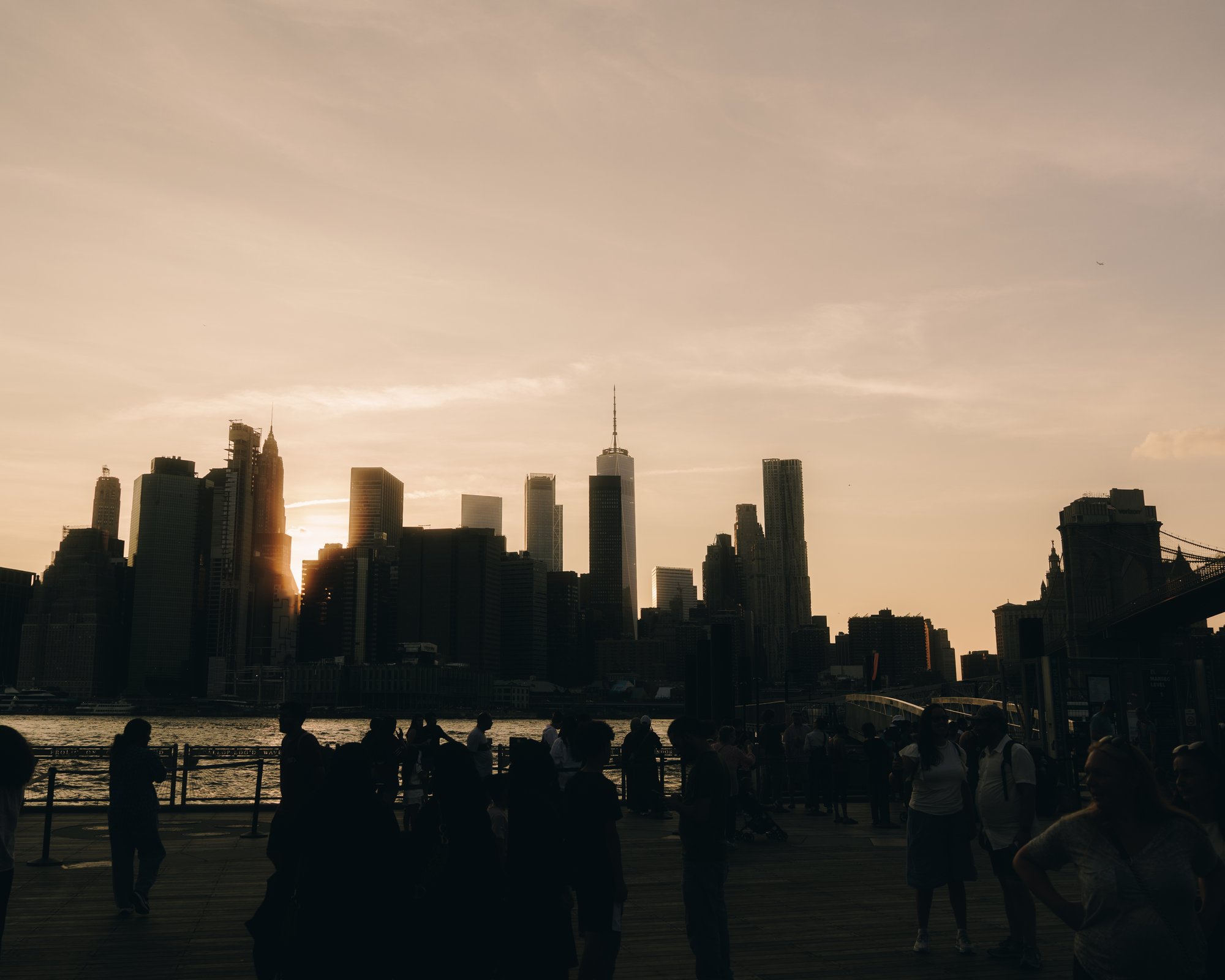 The Manhattan skyline and the Brooklyn Bridge at sunset, a crowd of onlookers along the promenade in silhouette