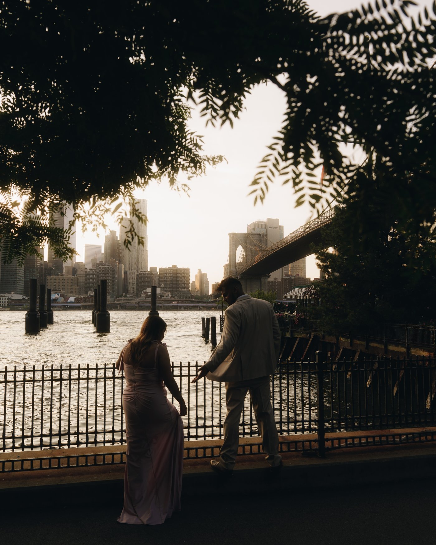 Seen through overhanging leaves, a couple at an iron railing above the water with the bridge and the skyline beyond