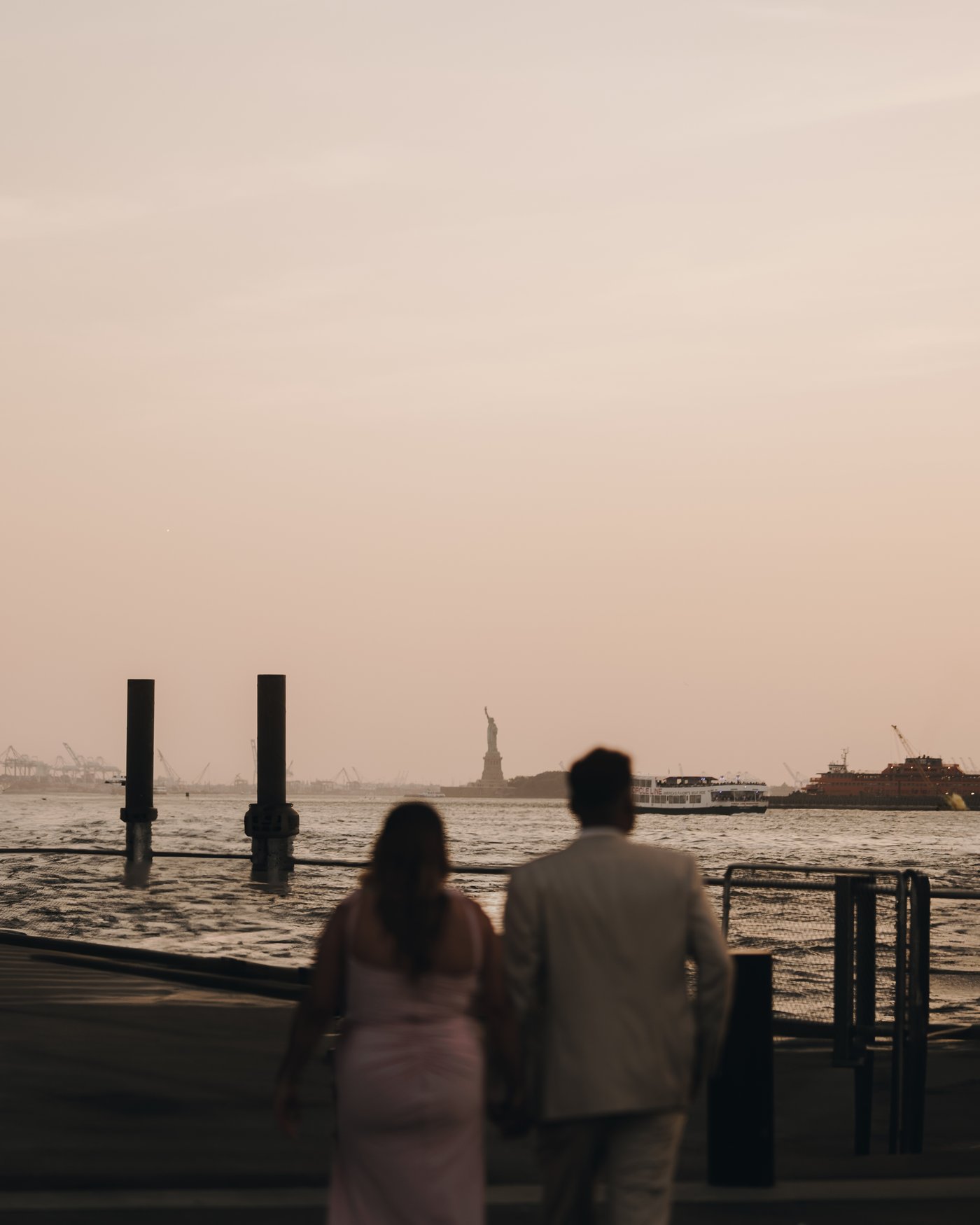 A couple from behind at the water's edge, holding hands and looking out toward the Statue of Liberty