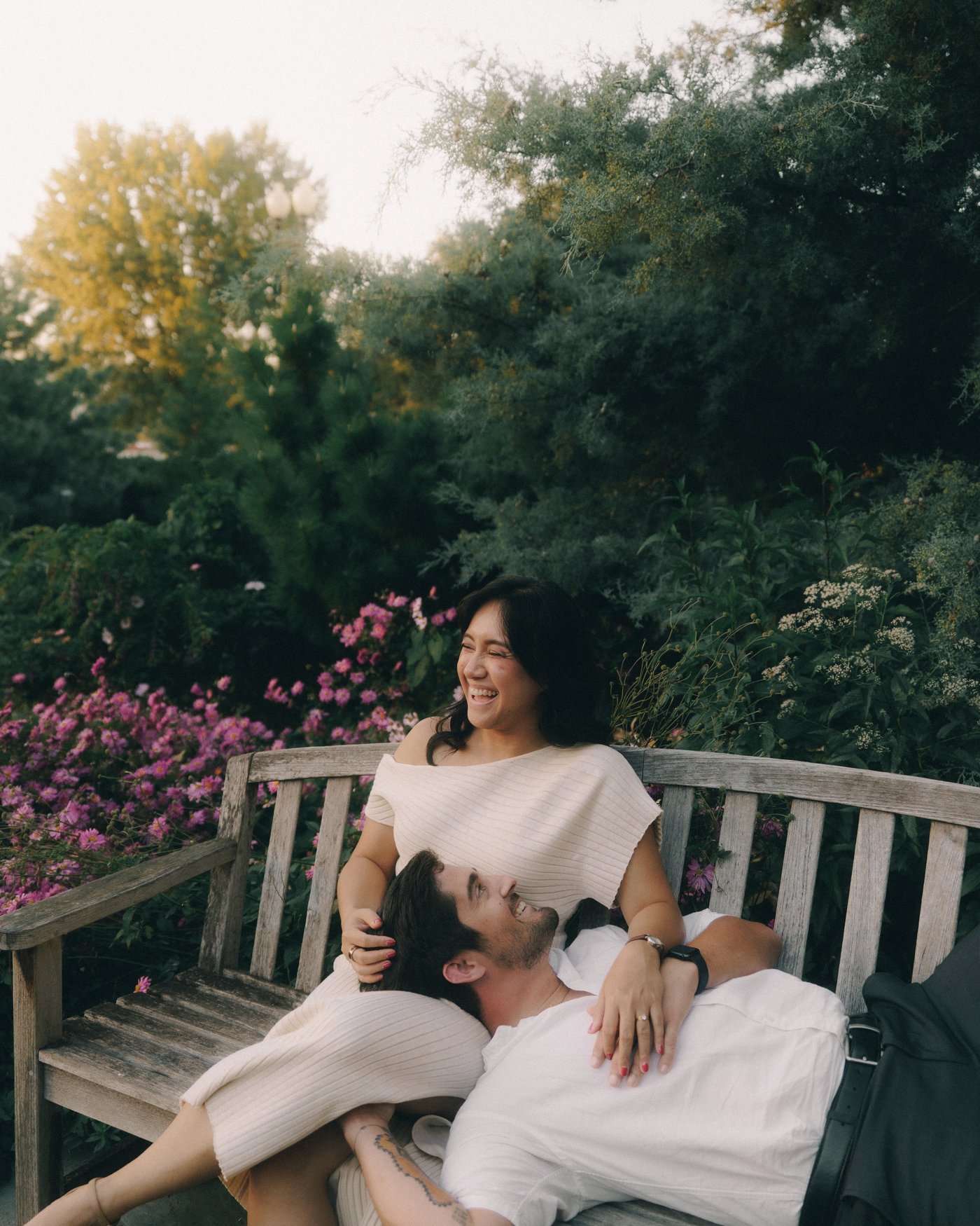 A woman laughing on a garden bench with her partner stretched back across her lap, roses behind them