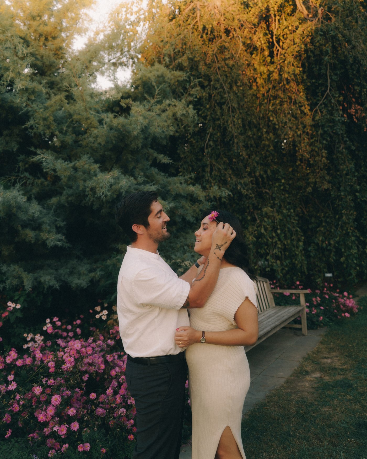 A man with a hand at his partner's cheek among flowering shrubs, the trees behind them turning gold