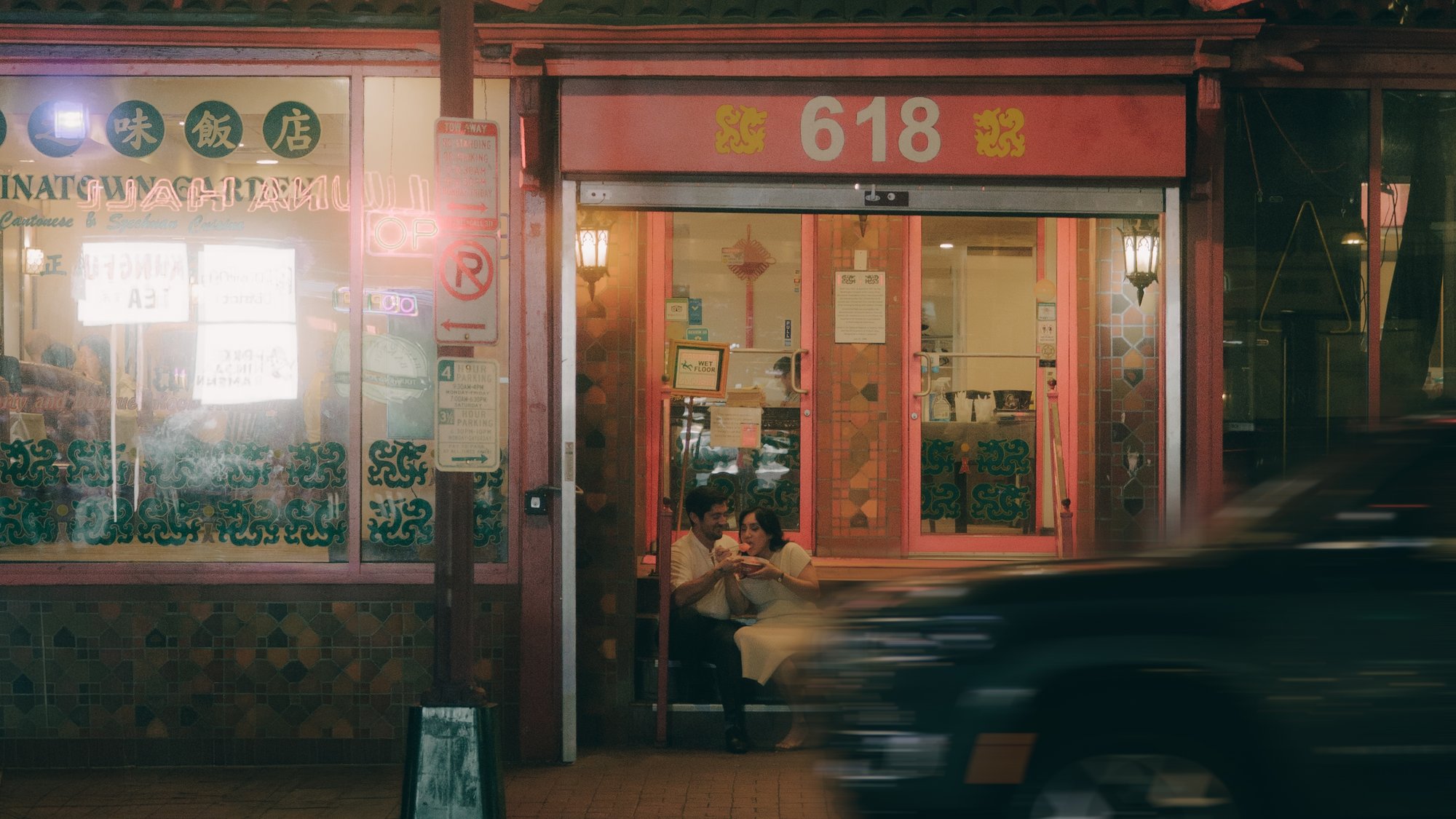 A Chinatown storefront at night, warm light spilling from the doorway, a car streaking past in the foreground