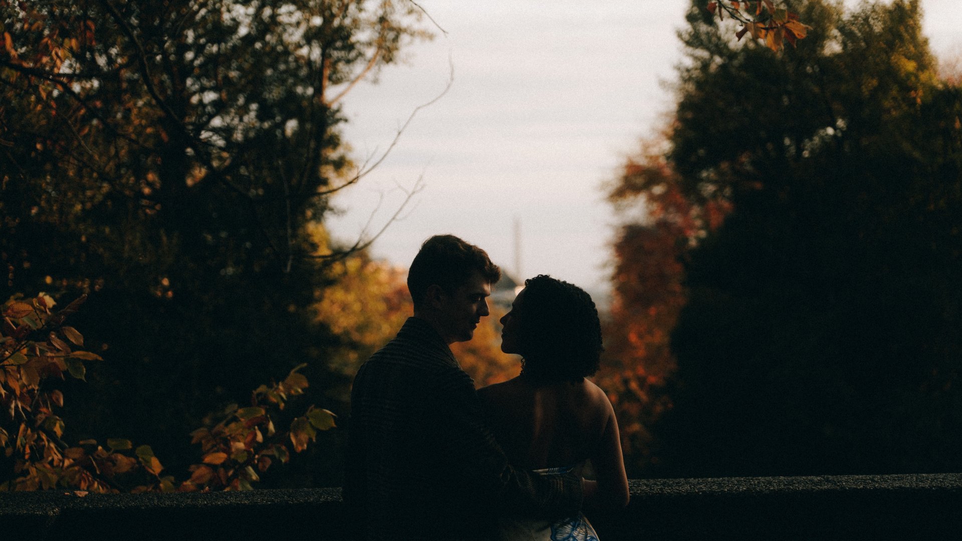A couple in silhouette facing each other at a stone parapet at dusk, autumn trees on both sides and the Washington Monument a pale needle on the horizon