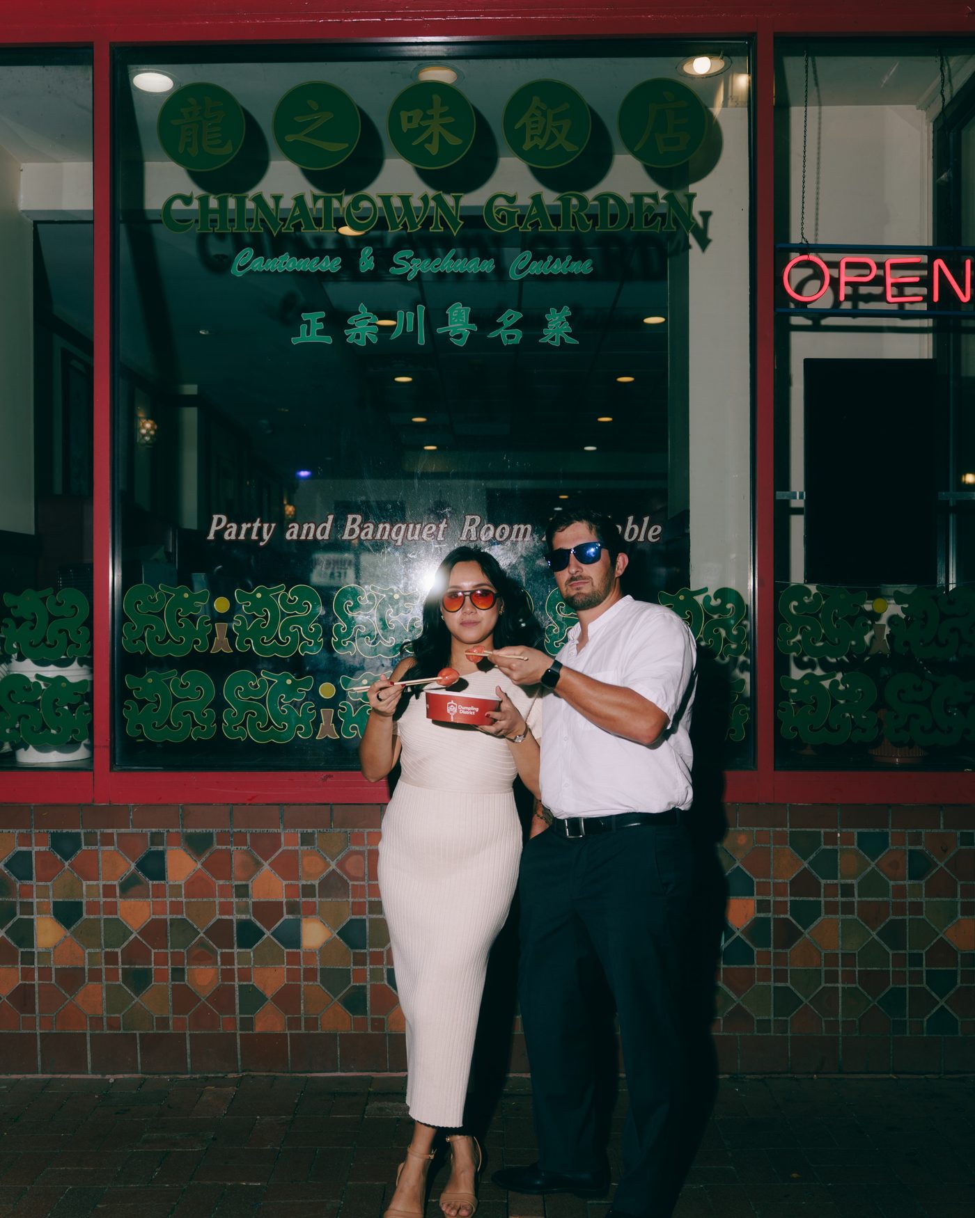 A couple in sunglasses outside a Chinatown restaurant, sharing a small red takeout box under the neon Open sign