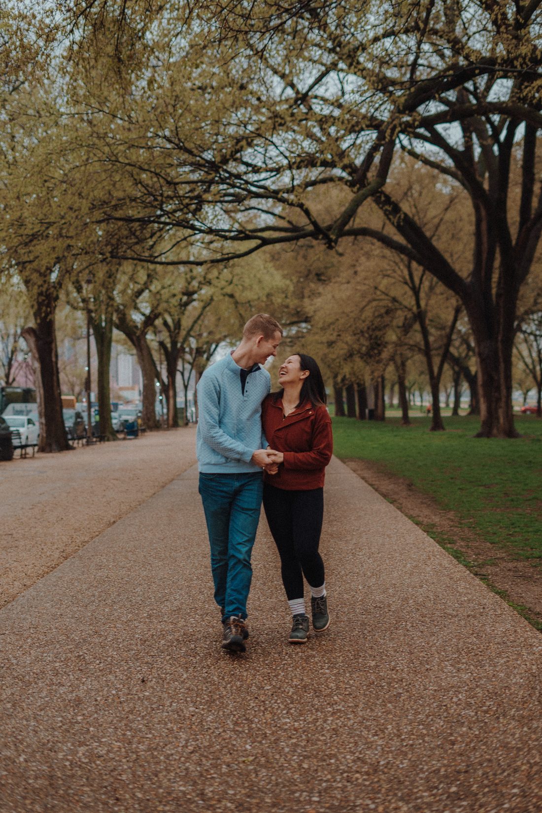 A couple walking arm in arm down a long gravel path under an avenue of trees