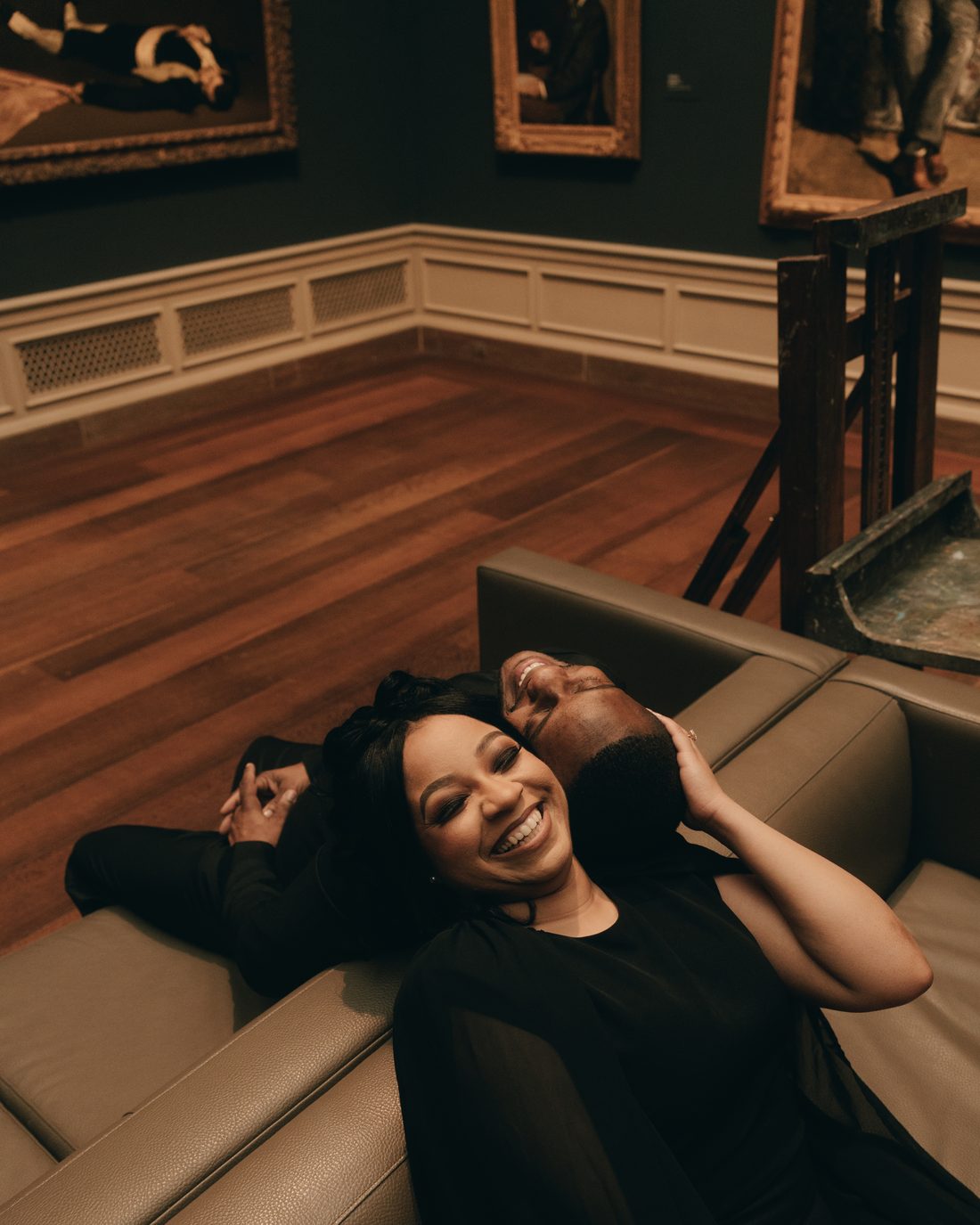 A couple lying back along a gallery bench, the woman laughing straight at the camera, gilt frames on the wall above
