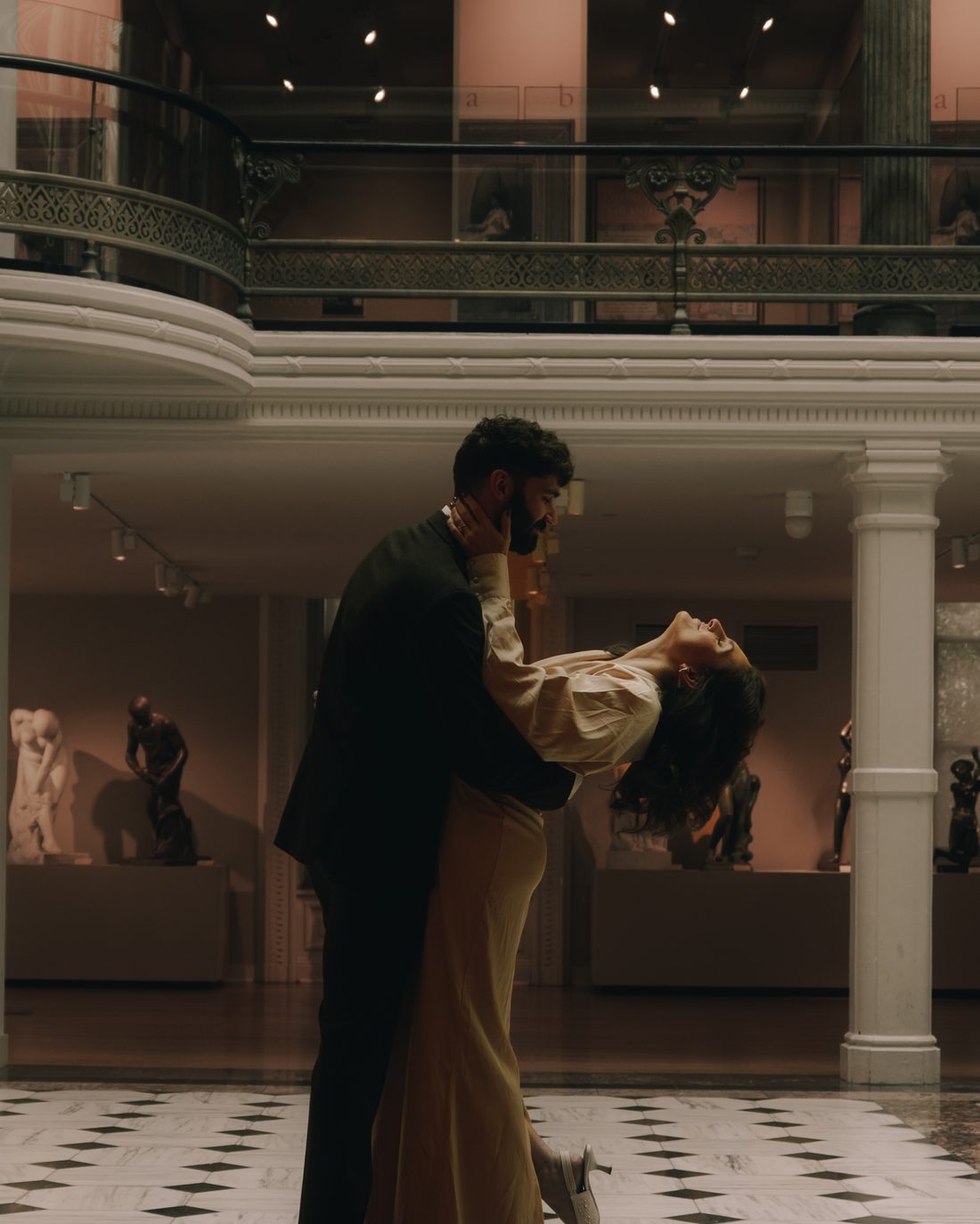 A man lifting his partner off her feet in a sculpture hall, marble checkerboard floor underneath
