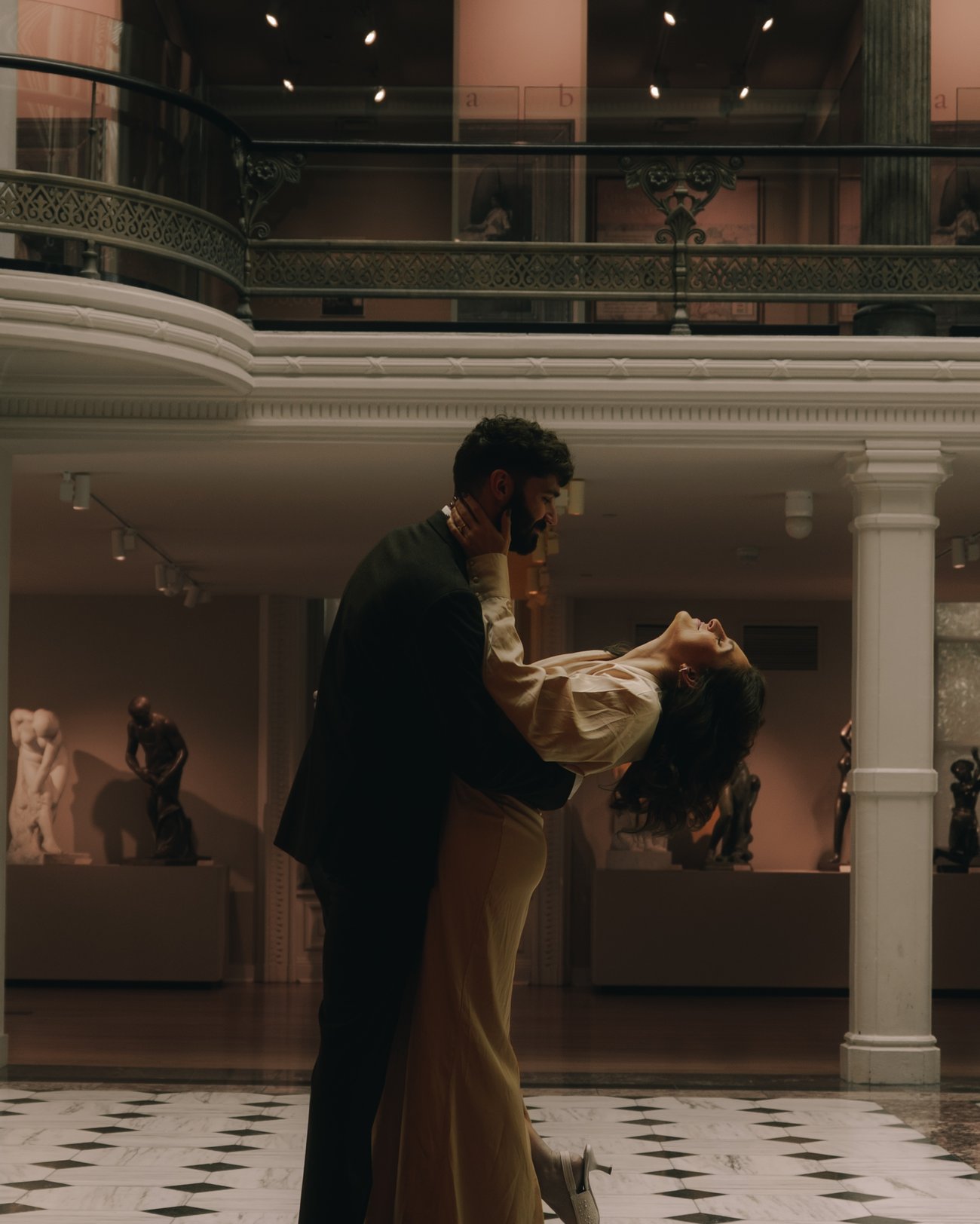 A man lifting his partner off her feet in a sculpture hall, marble checkerboard floor underneath