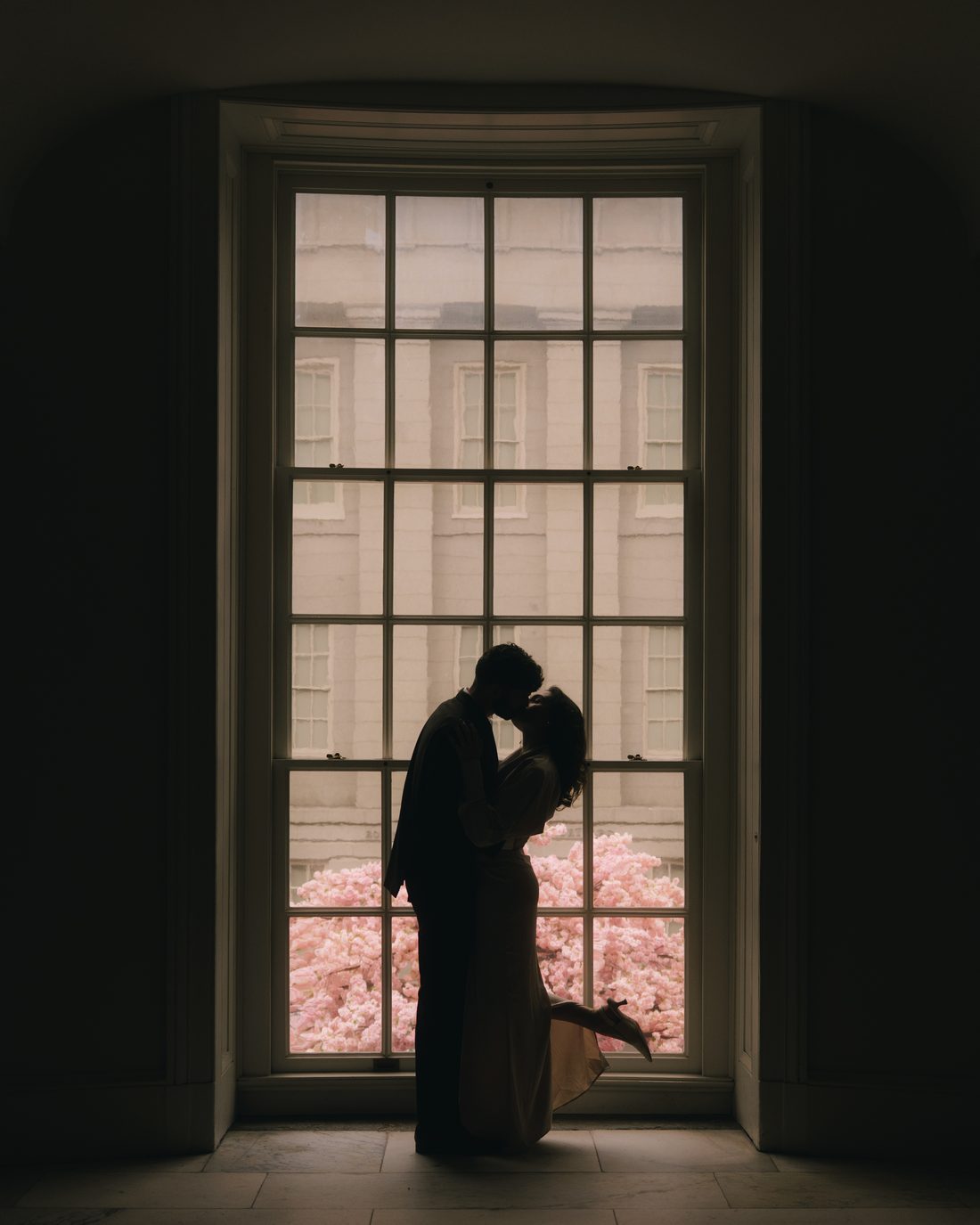 A couple silhouetted in a tall arched museum window with cherry blossom glowing pink outside it