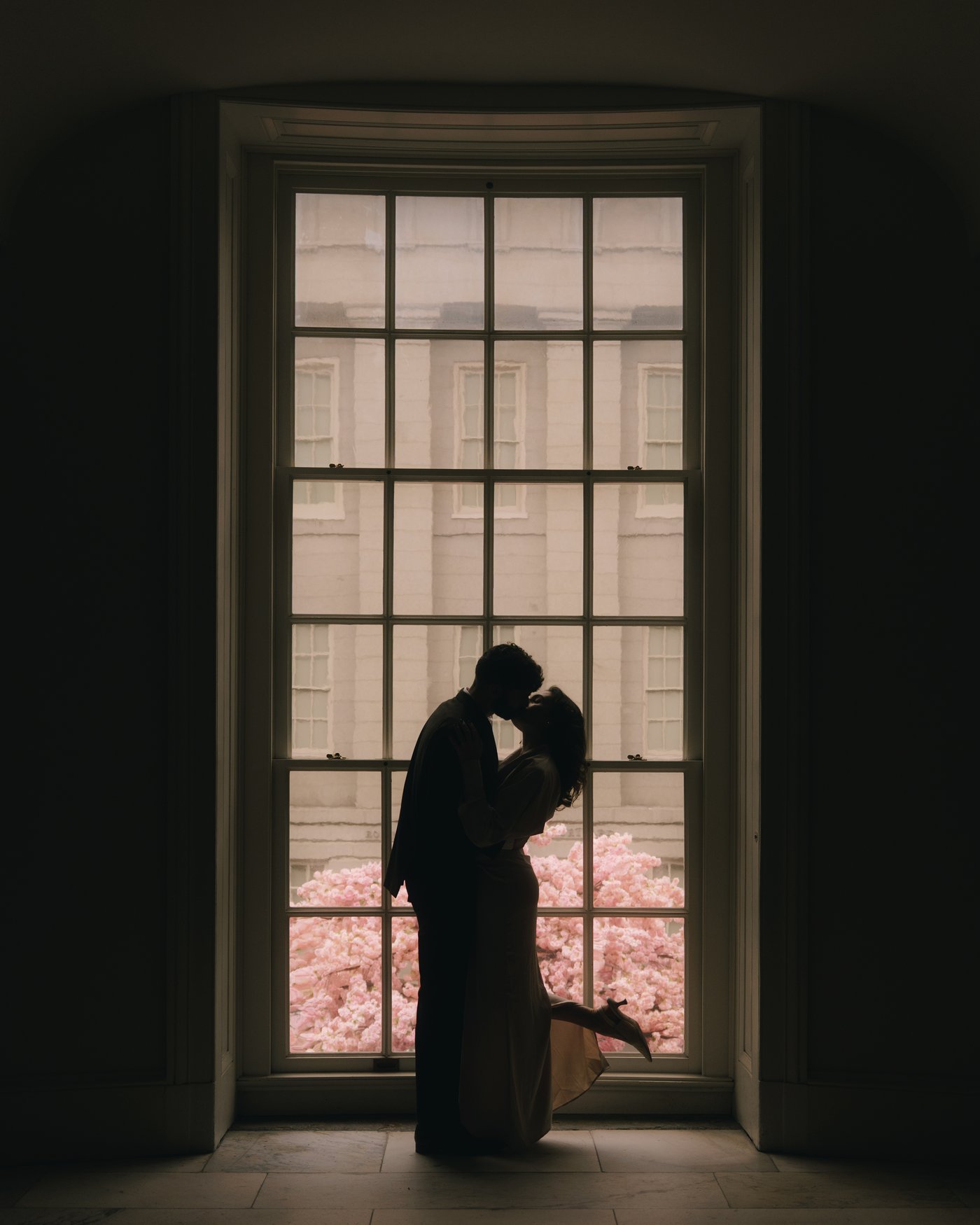 A couple silhouetted in a tall arched museum window with cherry blossom glowing pink outside it