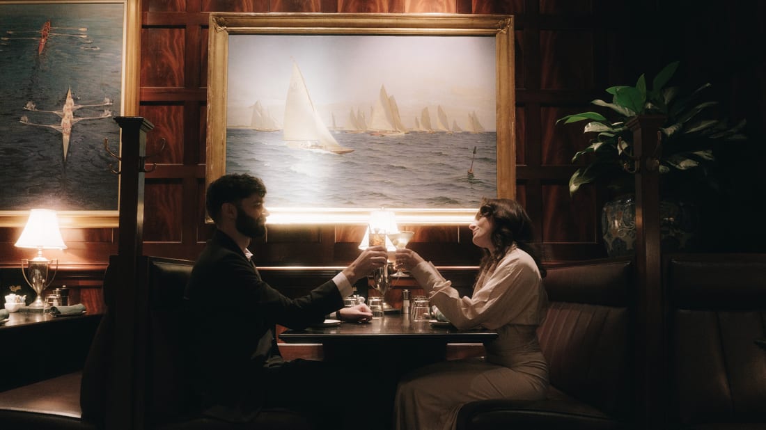 A wood panelled bar, a couple clinking glasses in a booth beneath a lit painting of sailboats