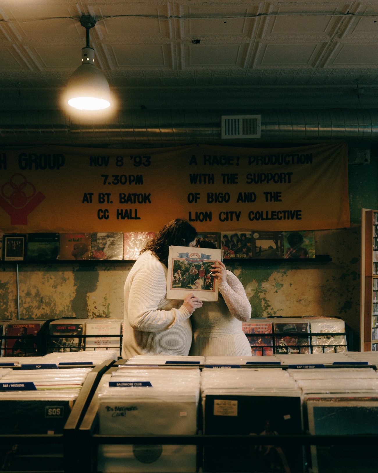 In a record shop, one of them holds an album sleeve up over their face while the other leans in behind the crates