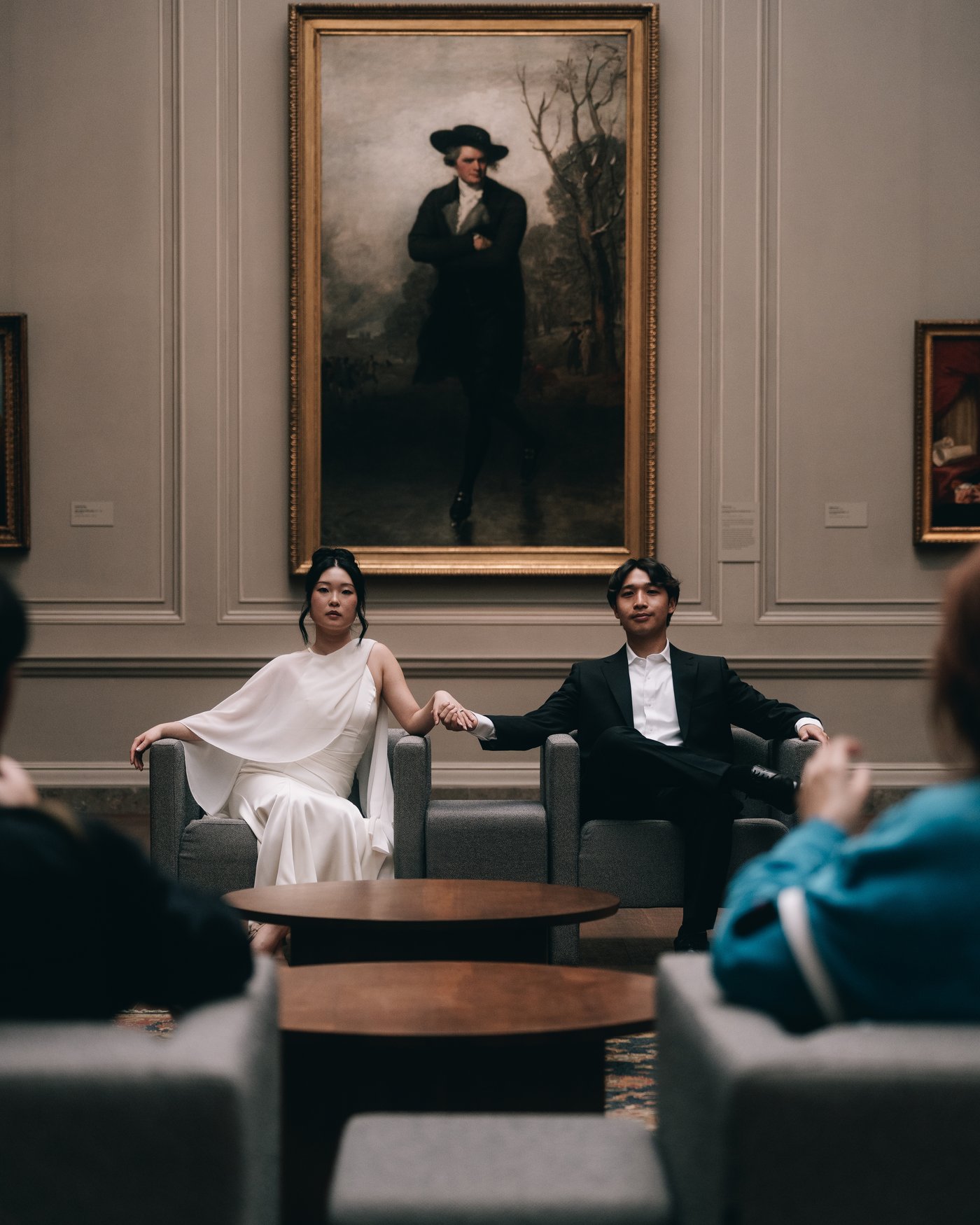 A couple holding hands across two armchairs in a museum lounge, a full length portrait on the wall behind them