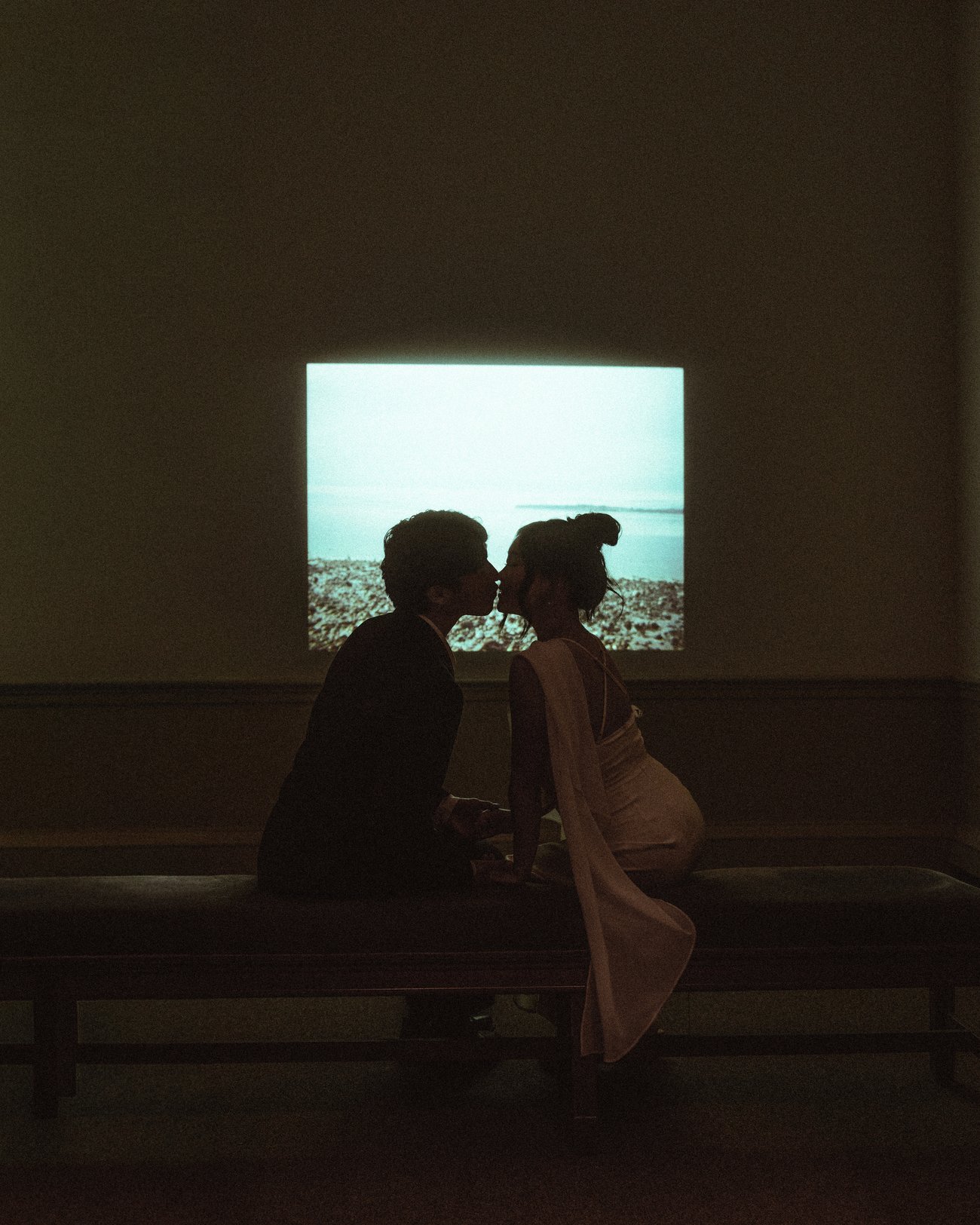 A couple close together on a bench in a dark room, lit only by the glow of a video projection on the wall