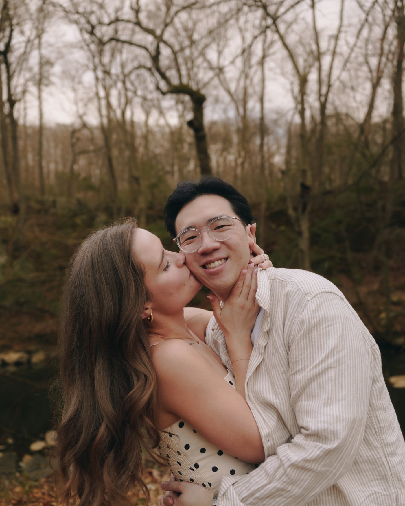 She kisses his cheek while he grins at the camera, bare branches behind them