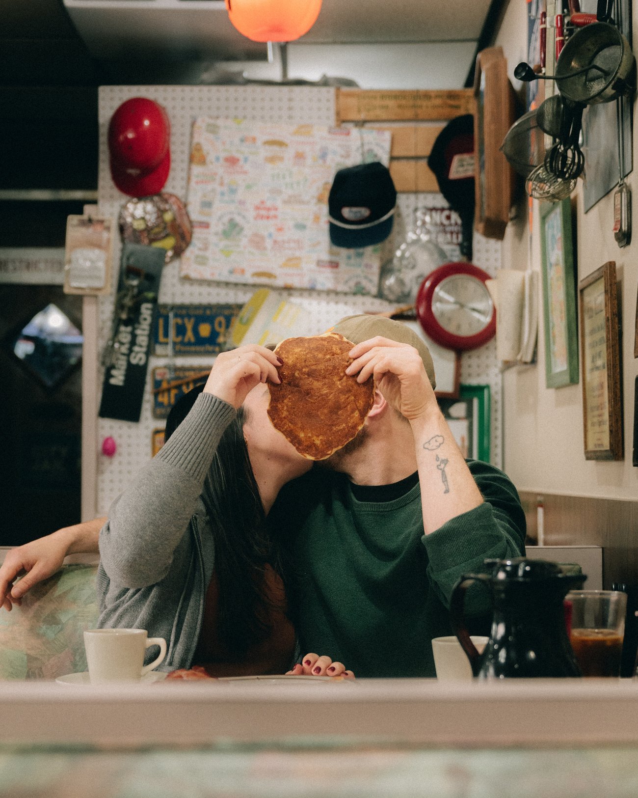In a cafe with every inch of wall pinned with notes and photographs, two people hiding behind an enormous cookie held up between their faces