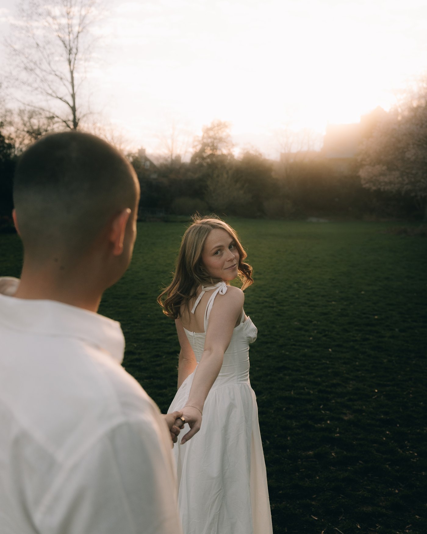 Over his shoulder, she turns back toward him across a dark field with the sun going down behind the treeline
