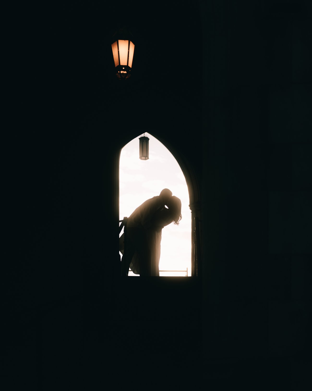 Two figures in a tight embrace framed by a dark stone archway, one lantern hanging above them