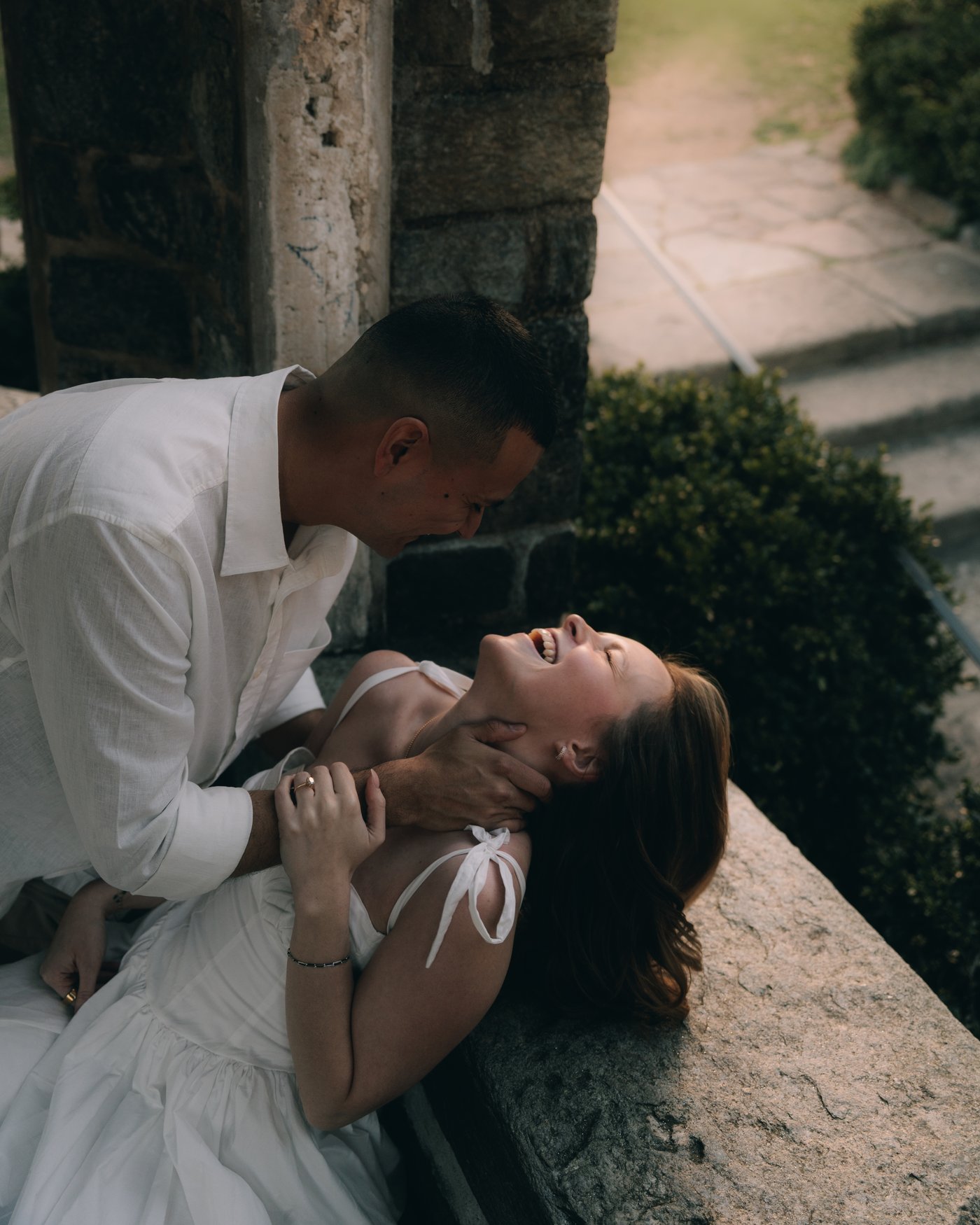 He leans over her as she lies back laughing along a stone ledge above the garden path