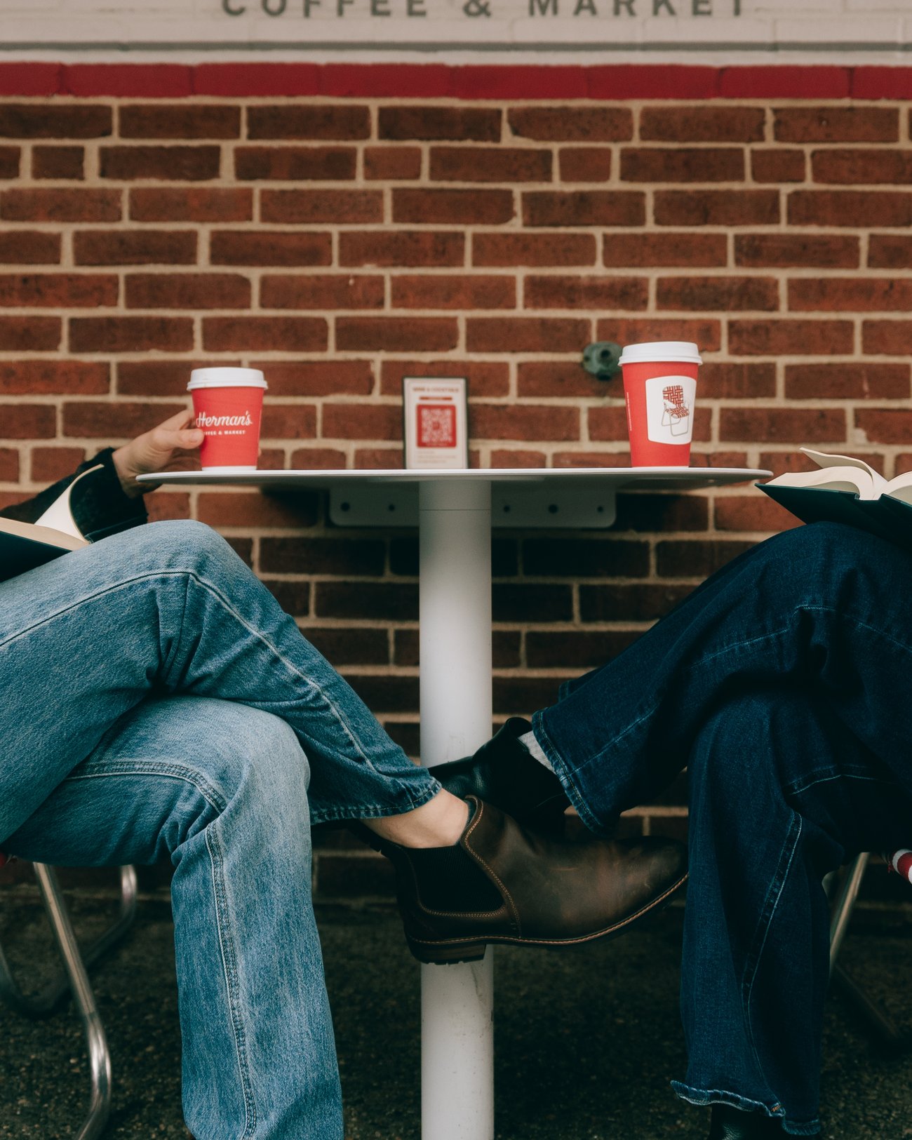 Cropped low at an outdoor cafe table: jeans, a boot hooked over the other chair, two red coffee cups on the tabletop above