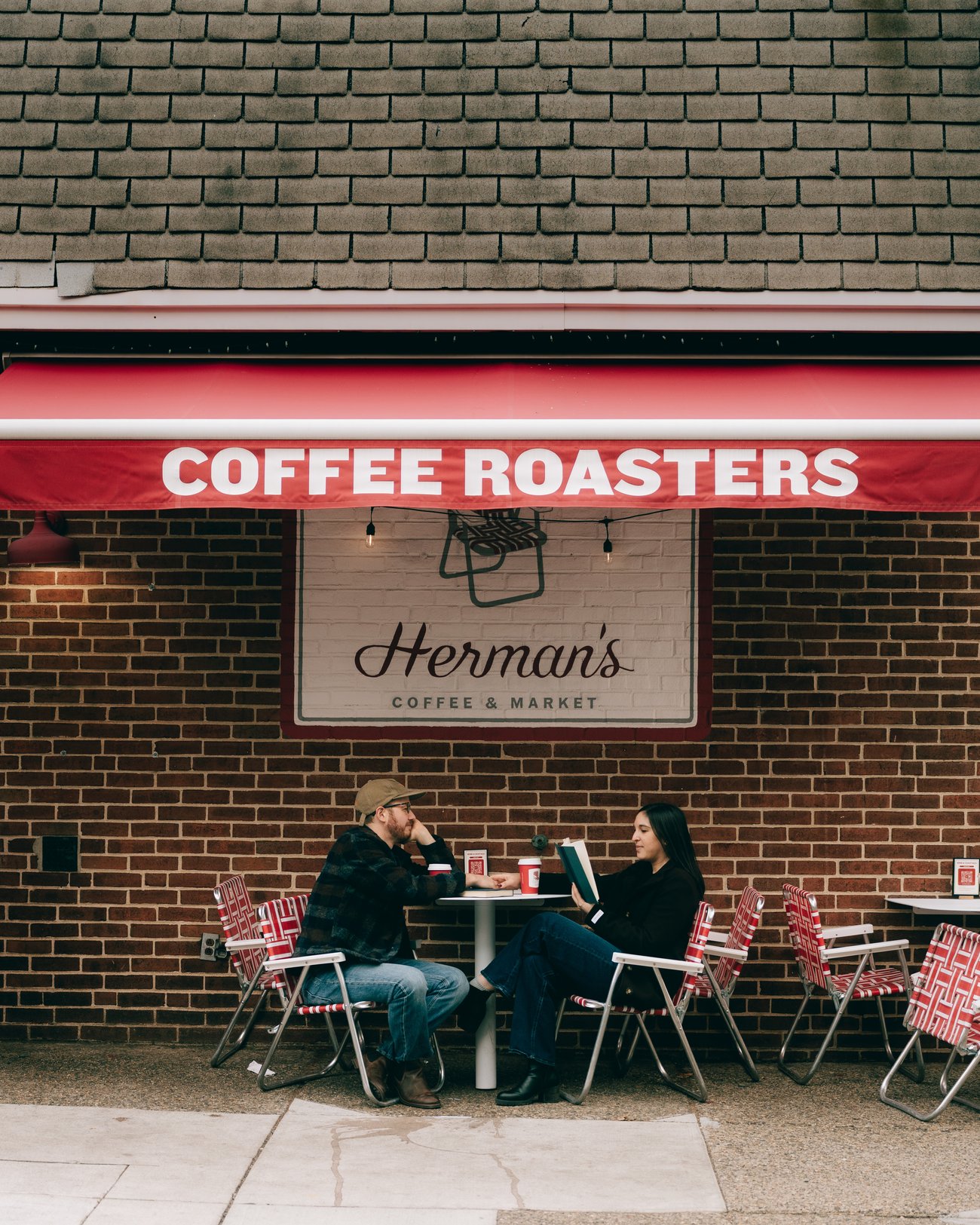 A red awninged coffee roaster seen from across the sidewalk, the couple at a small table out front in striped folding chairs