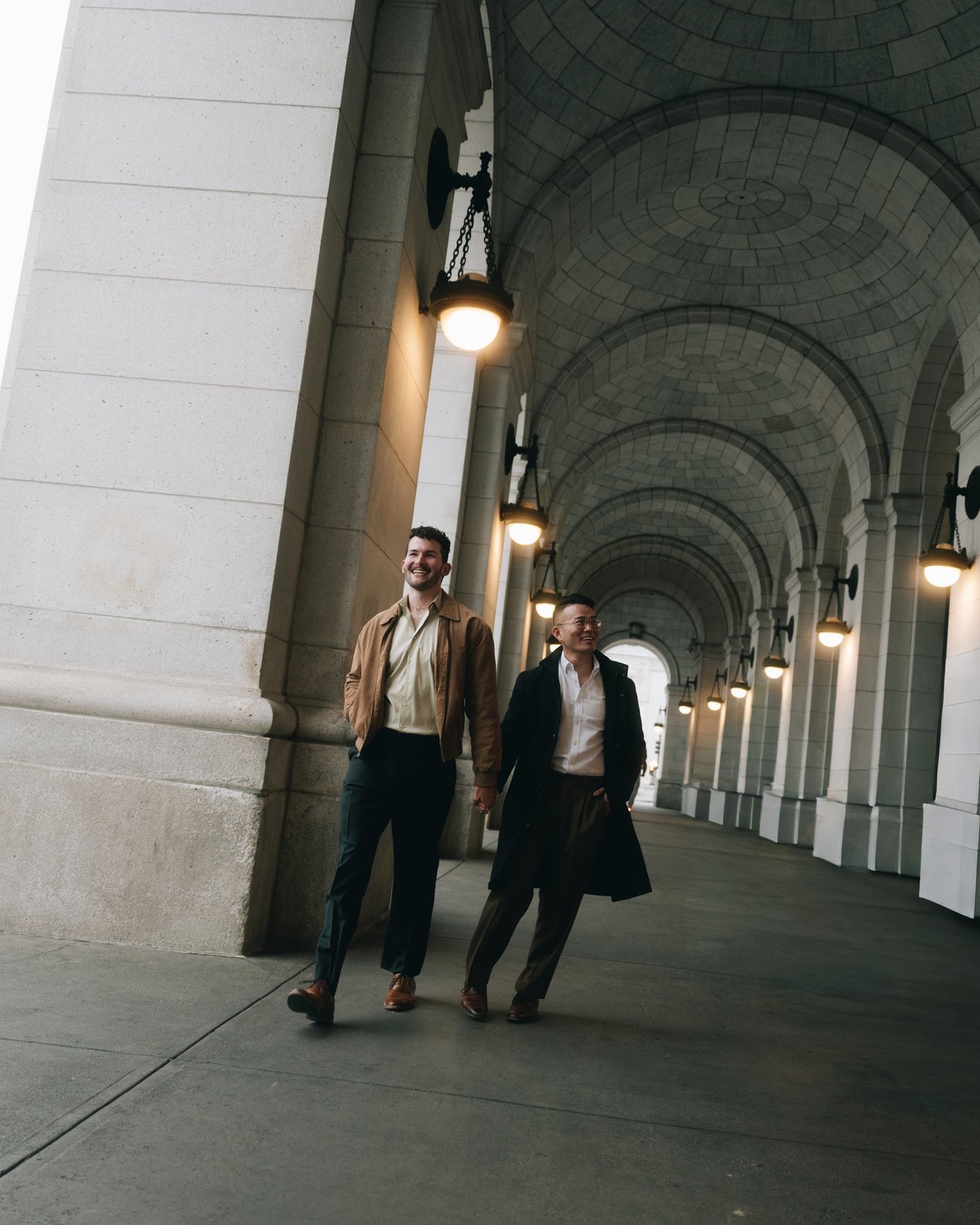 A couple walking hand in hand through a long vaulted stone arcade under hanging lamps