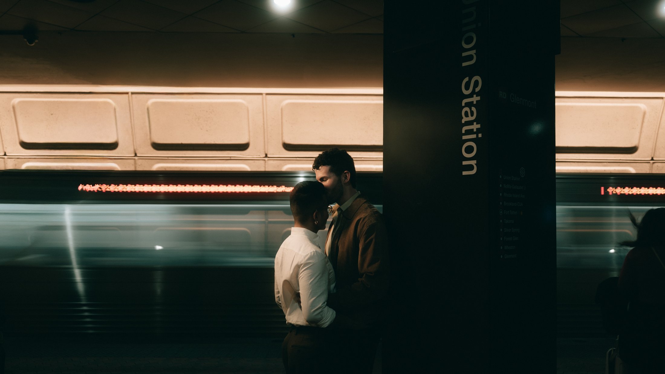 A train streaking past the platform at Union Station while a couple stand still together beside the sign