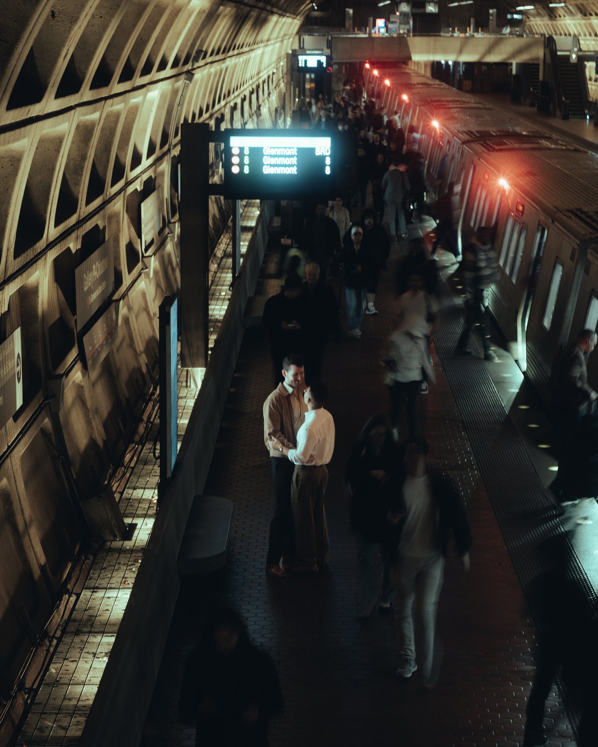 From above, a couple standing still and close together on a Metro platform at Union Station while the evening crowd blurs past them