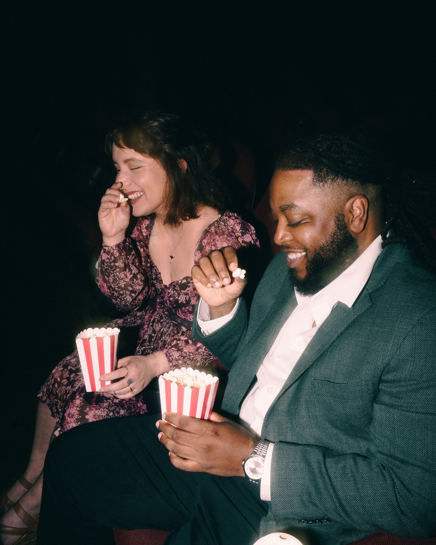 A couple in cinema seats with two boxes of popcorn, both mid laugh