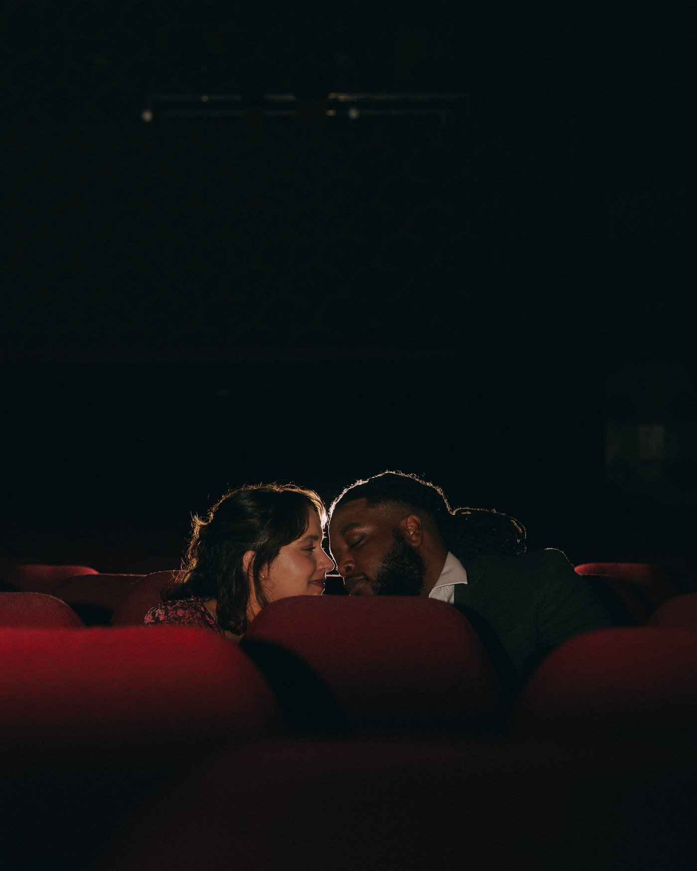 Foreheads together in the red velvet seats of a dark cinema, lit only by the screen