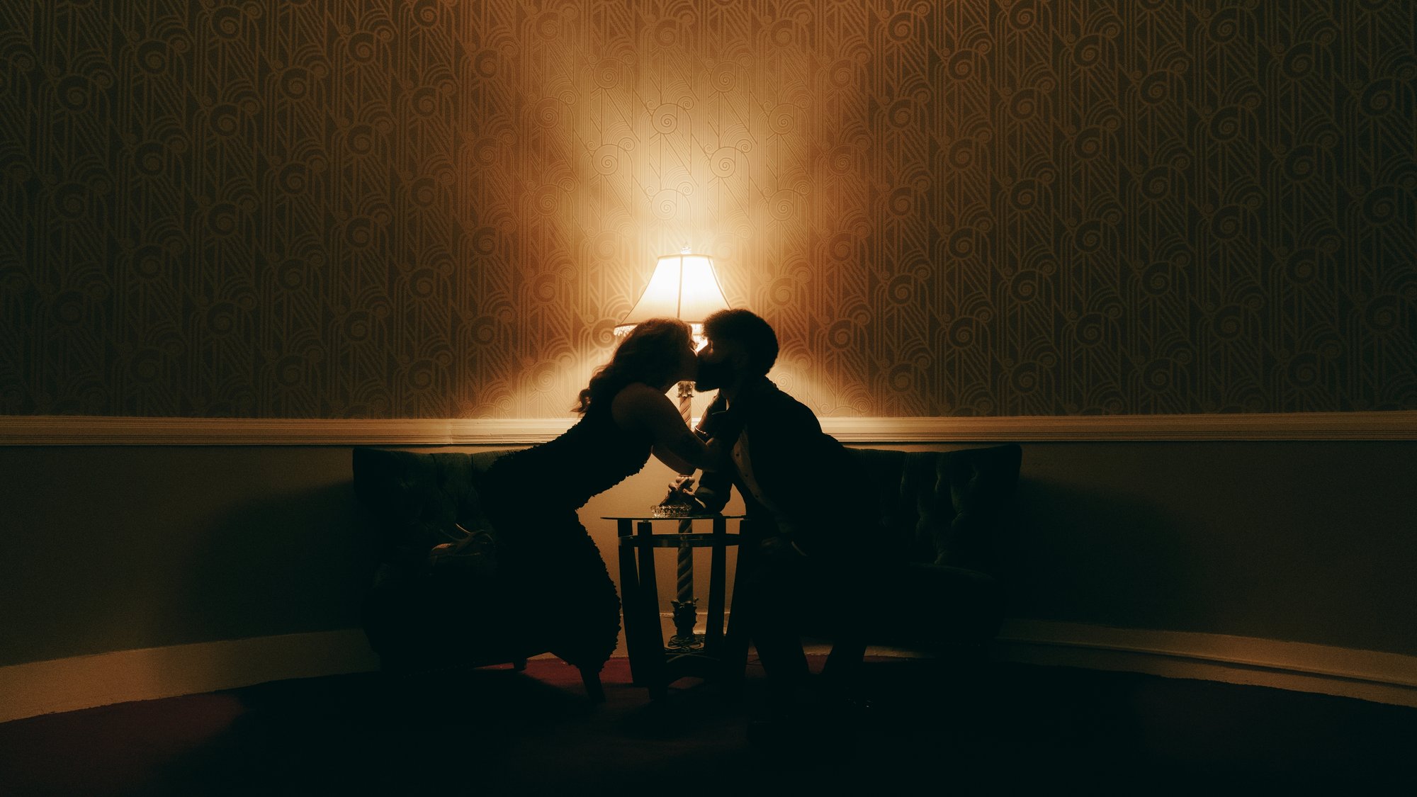 A dim wallpapered room lit by a single lamp, the couple leaning together into its glow