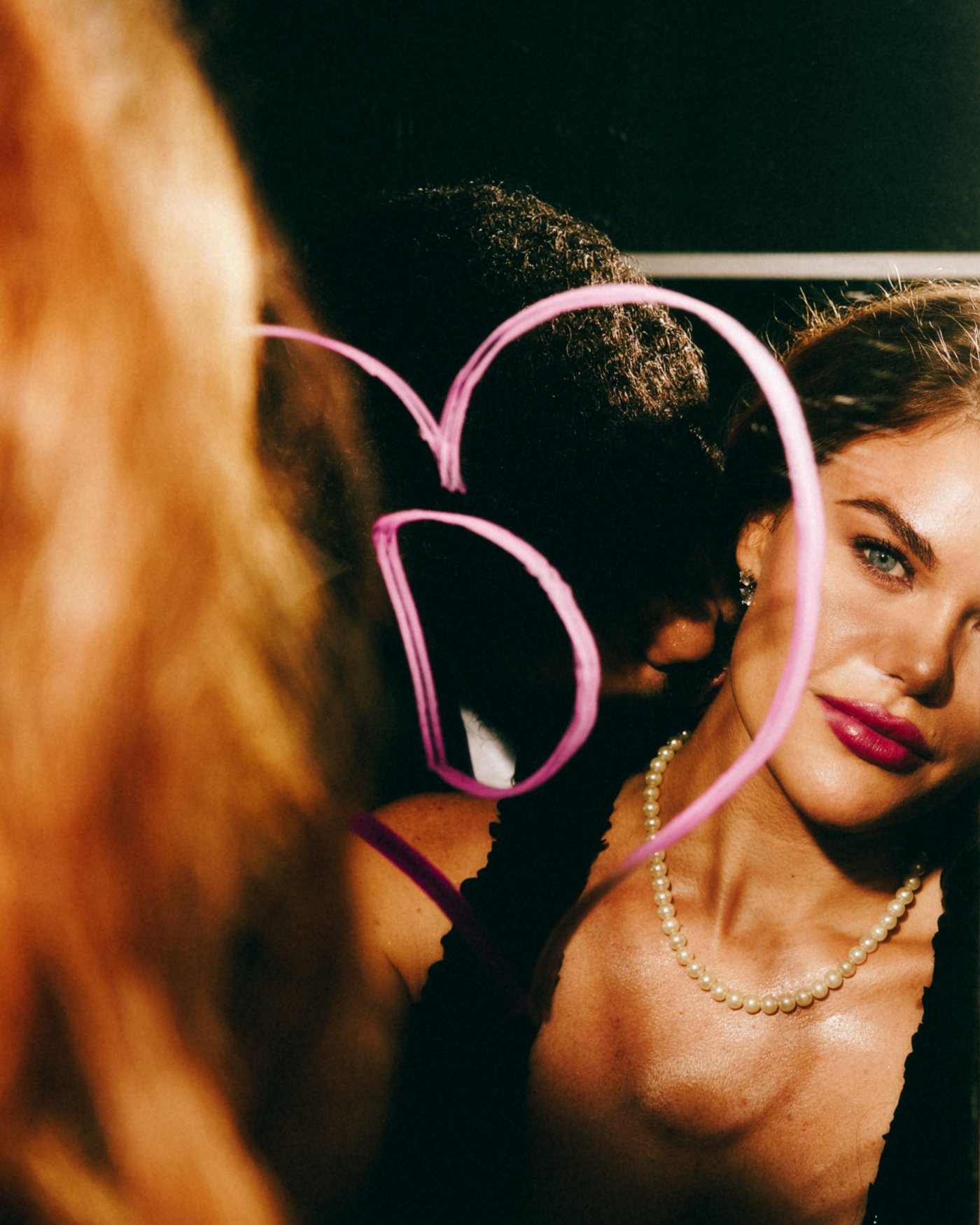 Close in at a party, a woman in pearls looking straight down the lens with a pink heart drawn in neon light hanging between her and her partner