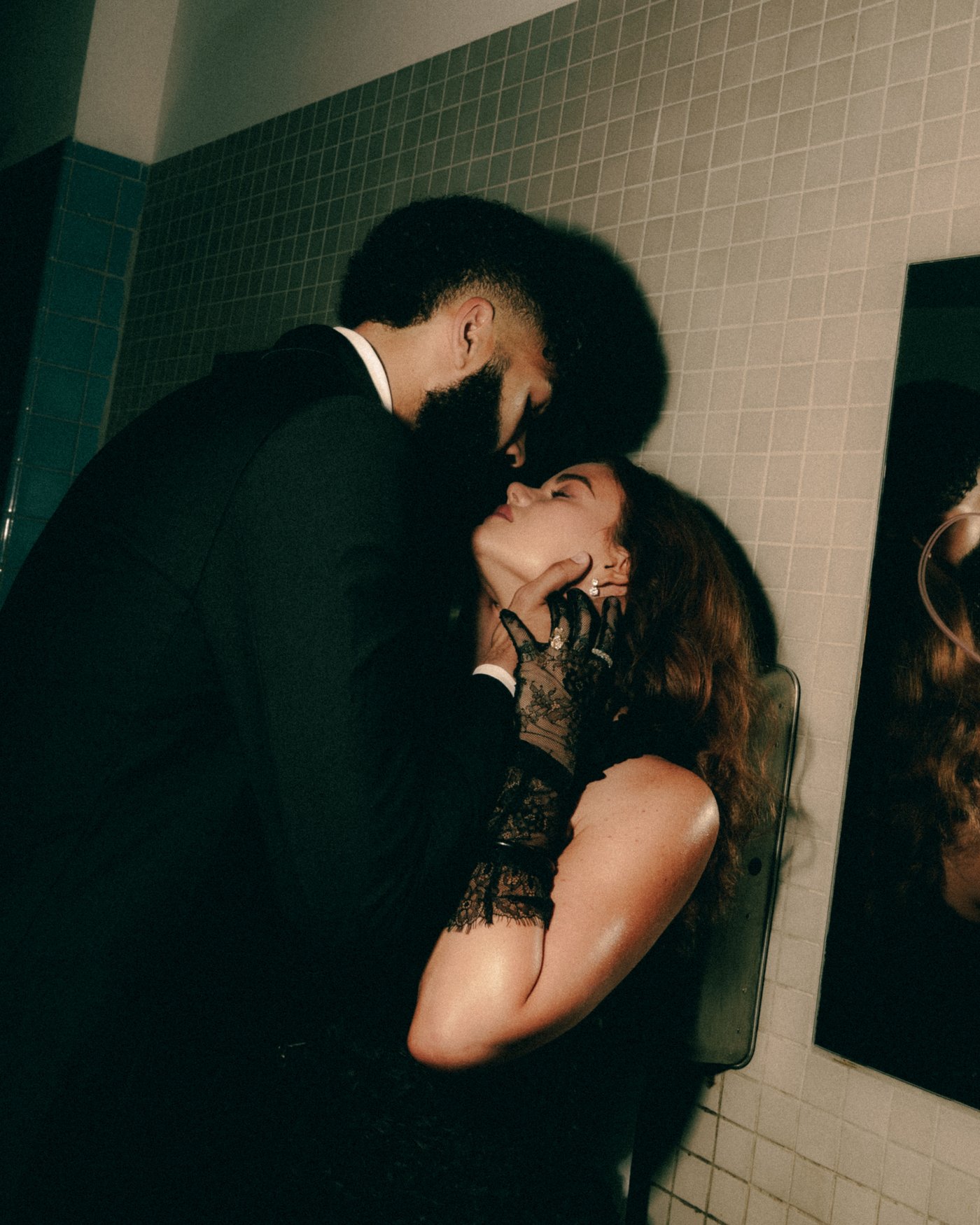 A couple kissing against a white tiled wall, her black lace sleeves catching the flash