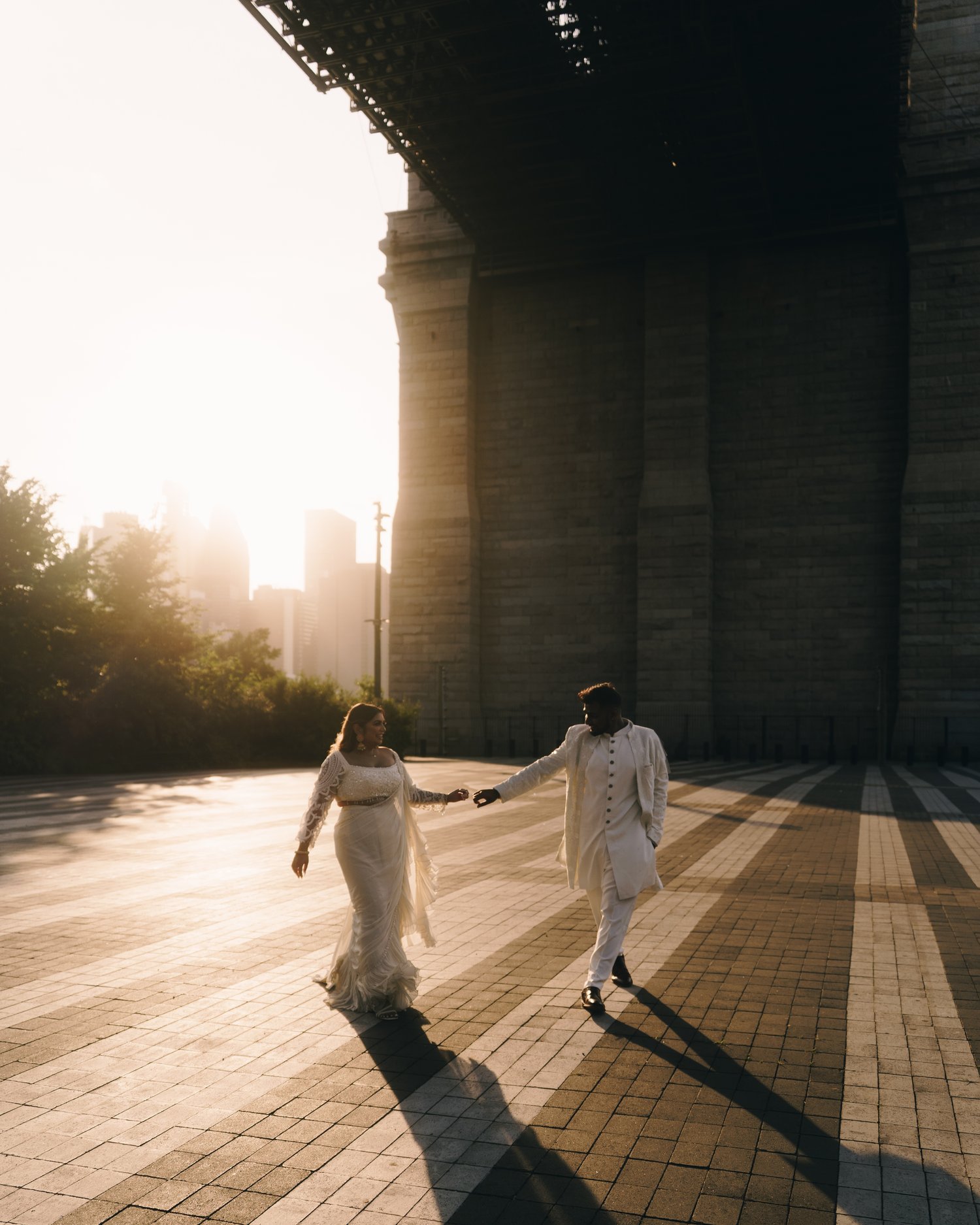 Two figures running hand in hand out of a bridge underpass into low sun, long shadows across the pavers