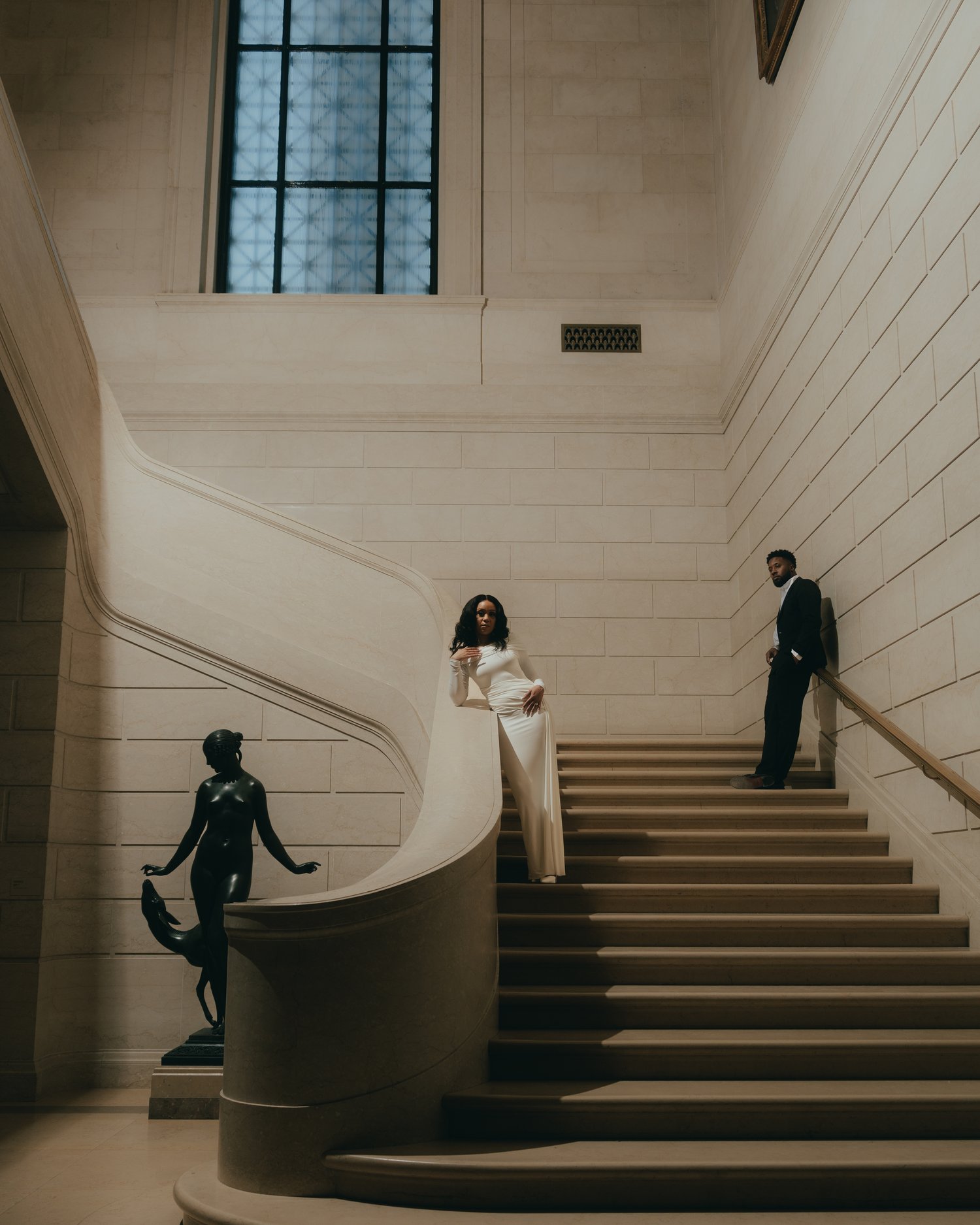 A pale marble stairwell with a bronze figure on its plinth, a woman in white on the landing and her partner on the steps above