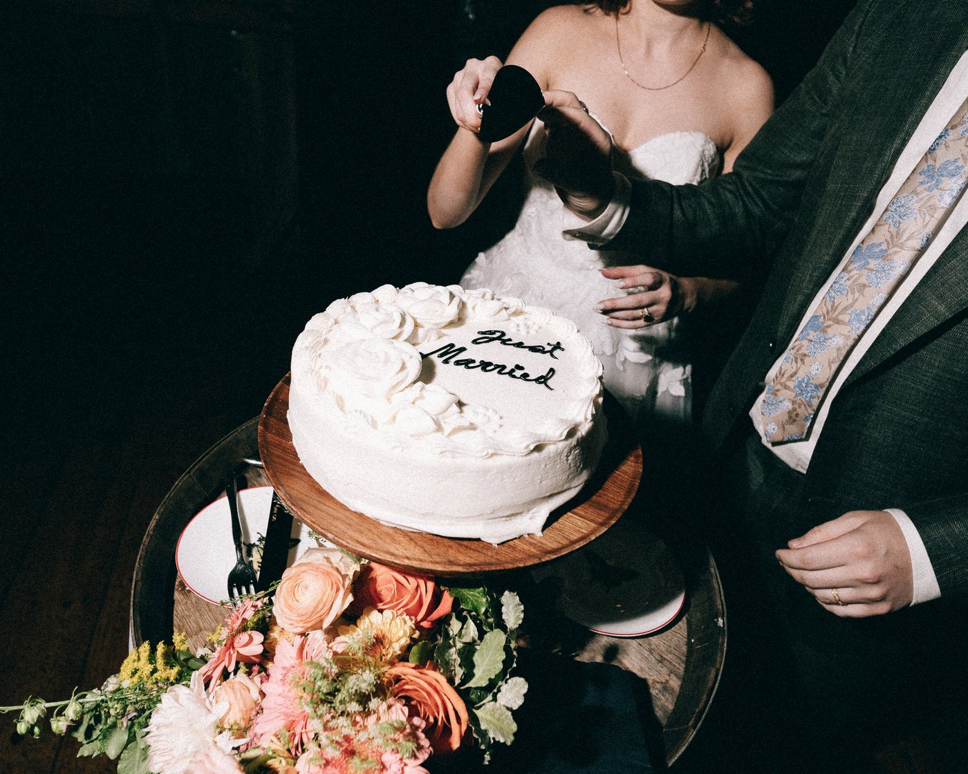 Hands cutting into the single tier wedding cake beside a bowl of flowers
