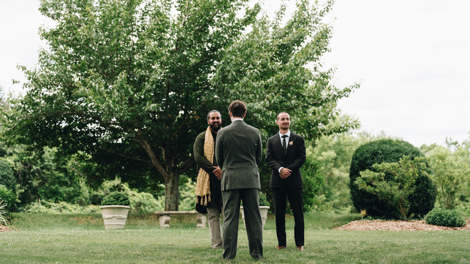 The groom waiting on the lawn with his back turned, two friends standing with him