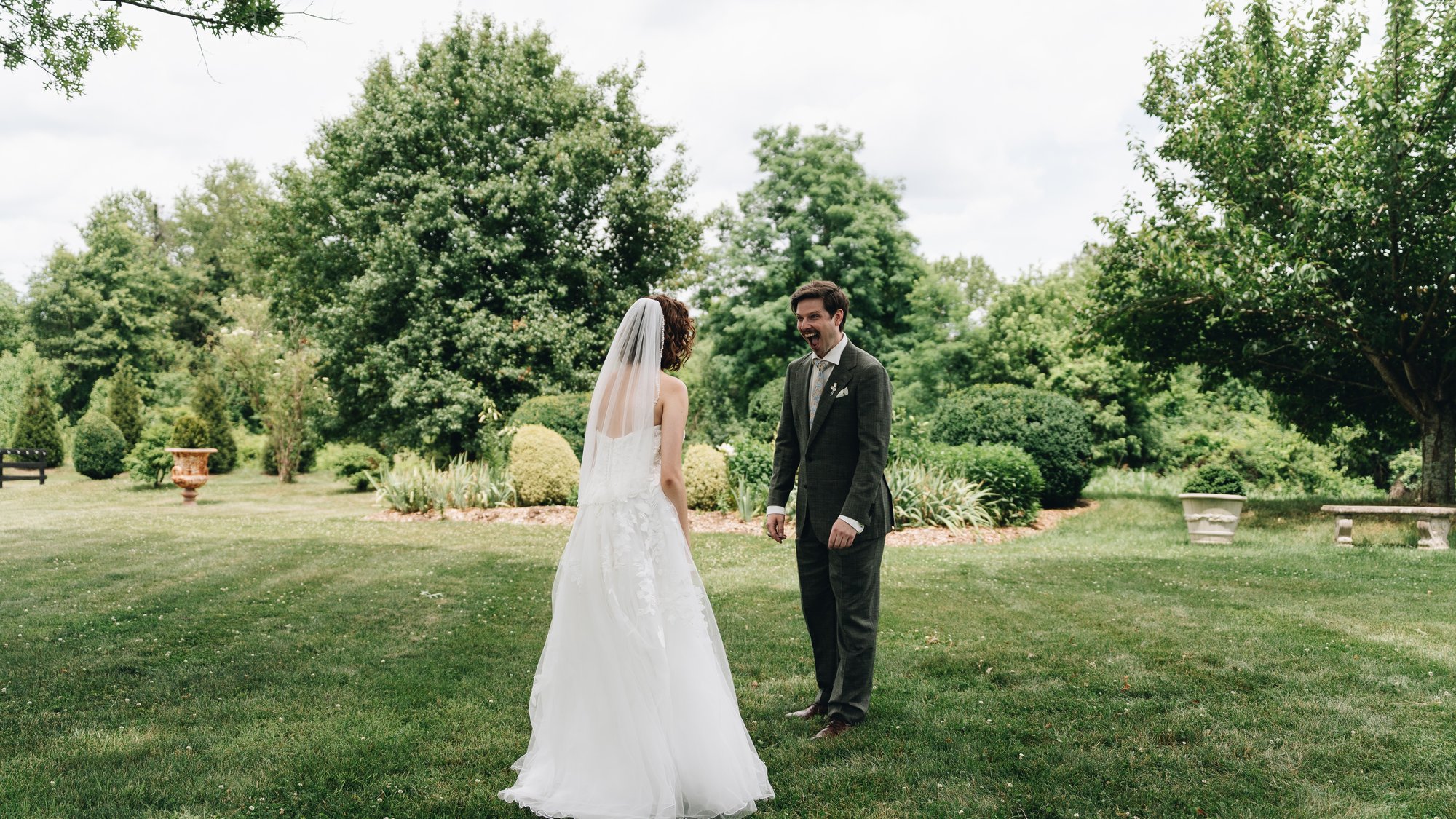 The bride and groom facing each other on the lawn at the first look, the groom laughing