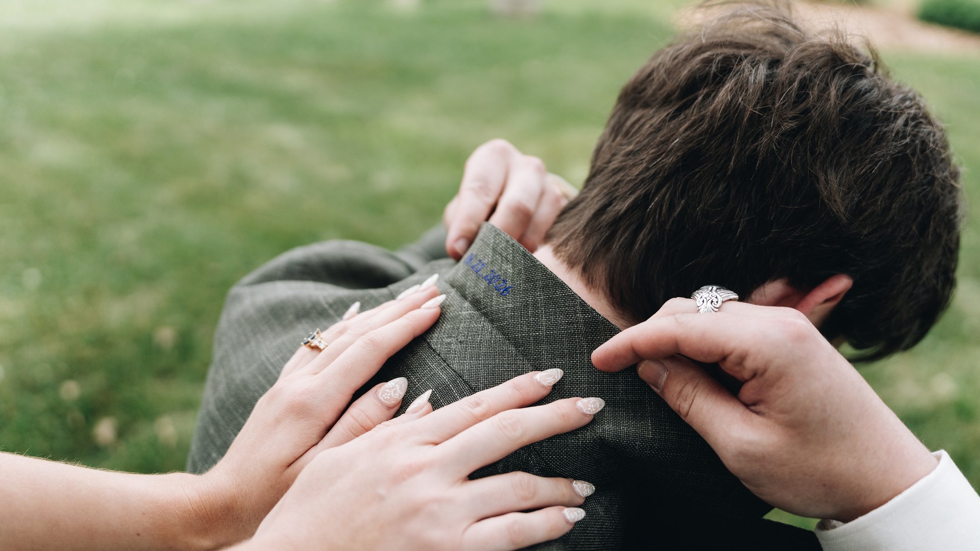 Close on the bride's hands folding back the groom's jacket collar, where he had had their wedding date embroidered in blue thread