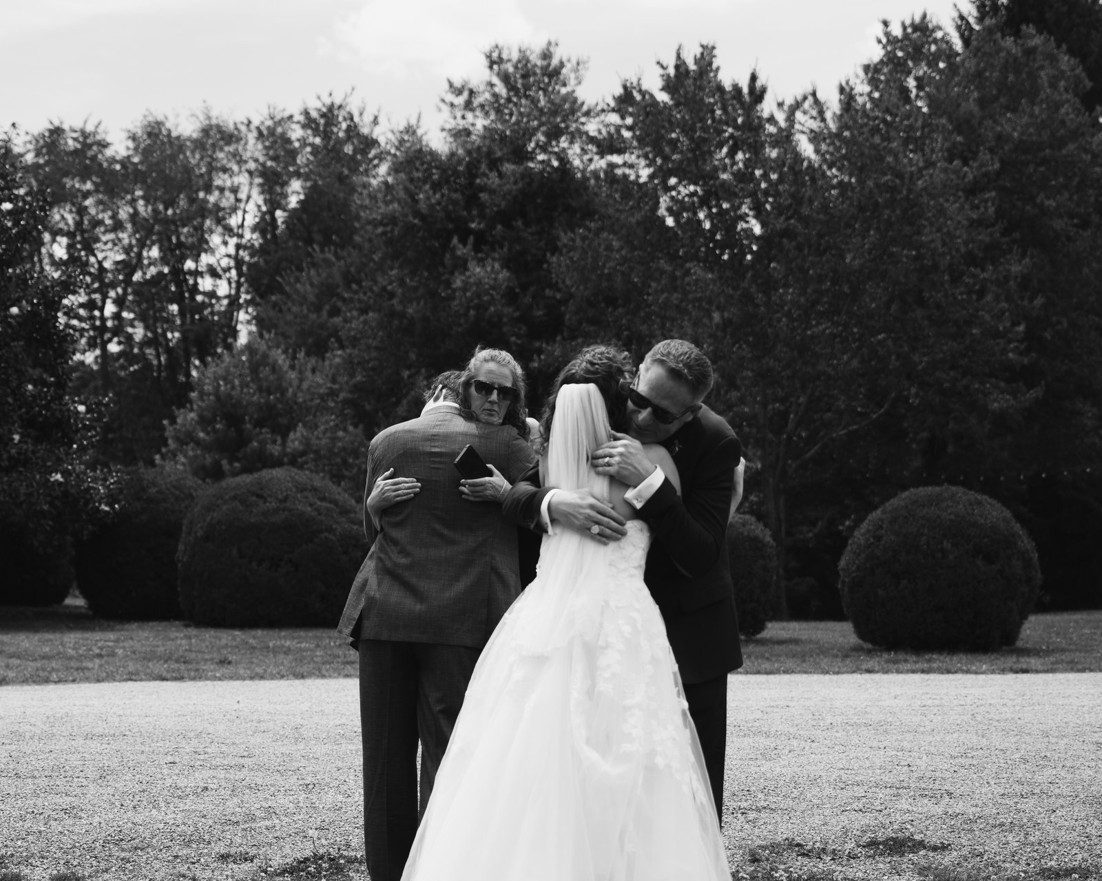Black and white, the bride held in a long embrace by two men in dark suits and sunglasses