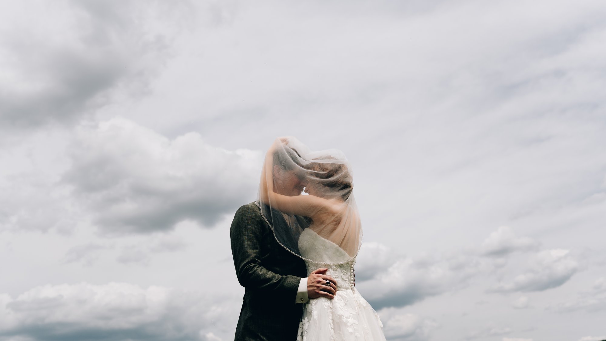 The bride's veil blowing across the couple as they kiss against a bank of clouds