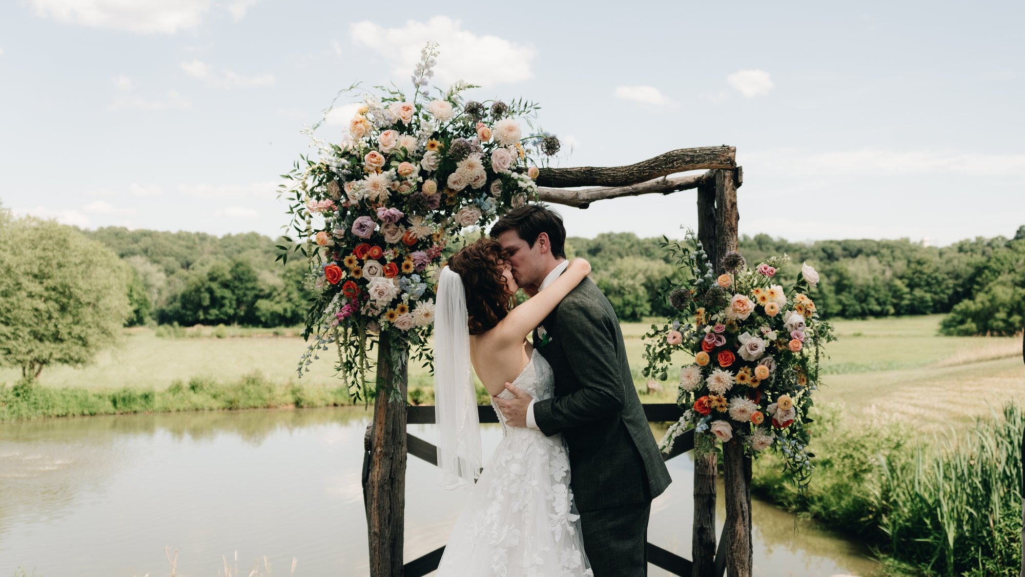 The bride and groom kissing under the floral arch with the pond behind them