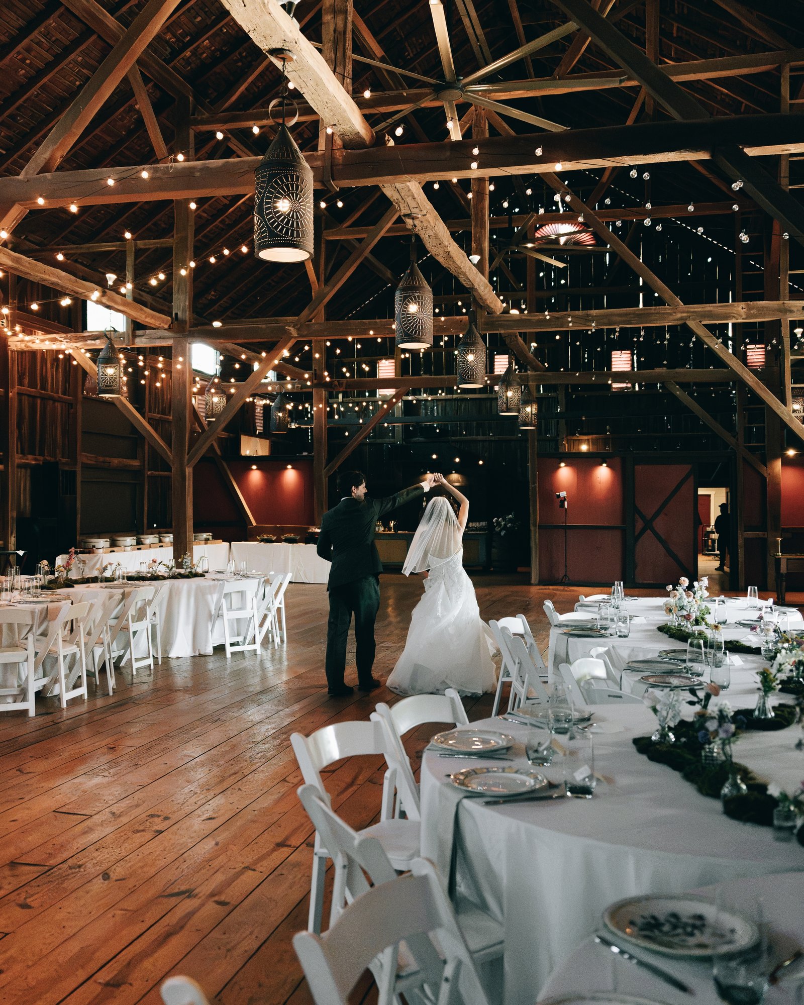 The barn dining room laid with round tables under string lights, the couple dancing in the middle of it