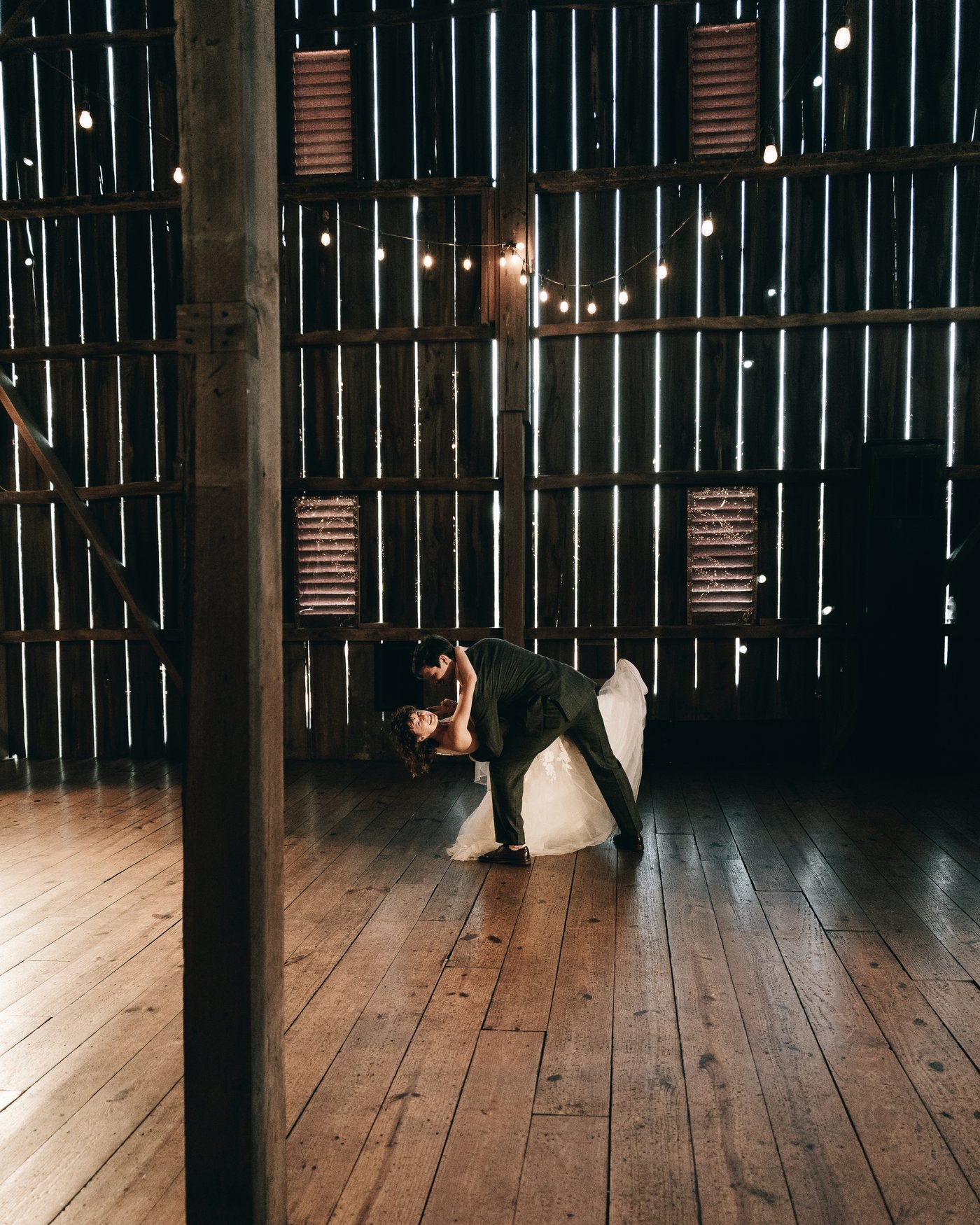 The groom dipping the bride on the barn's wooden floor, a timber post in the foreground