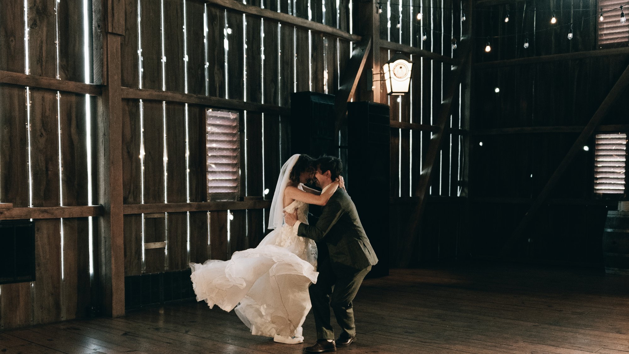 The bride and groom dancing against the slatted barn wall, daylight coming through the boards