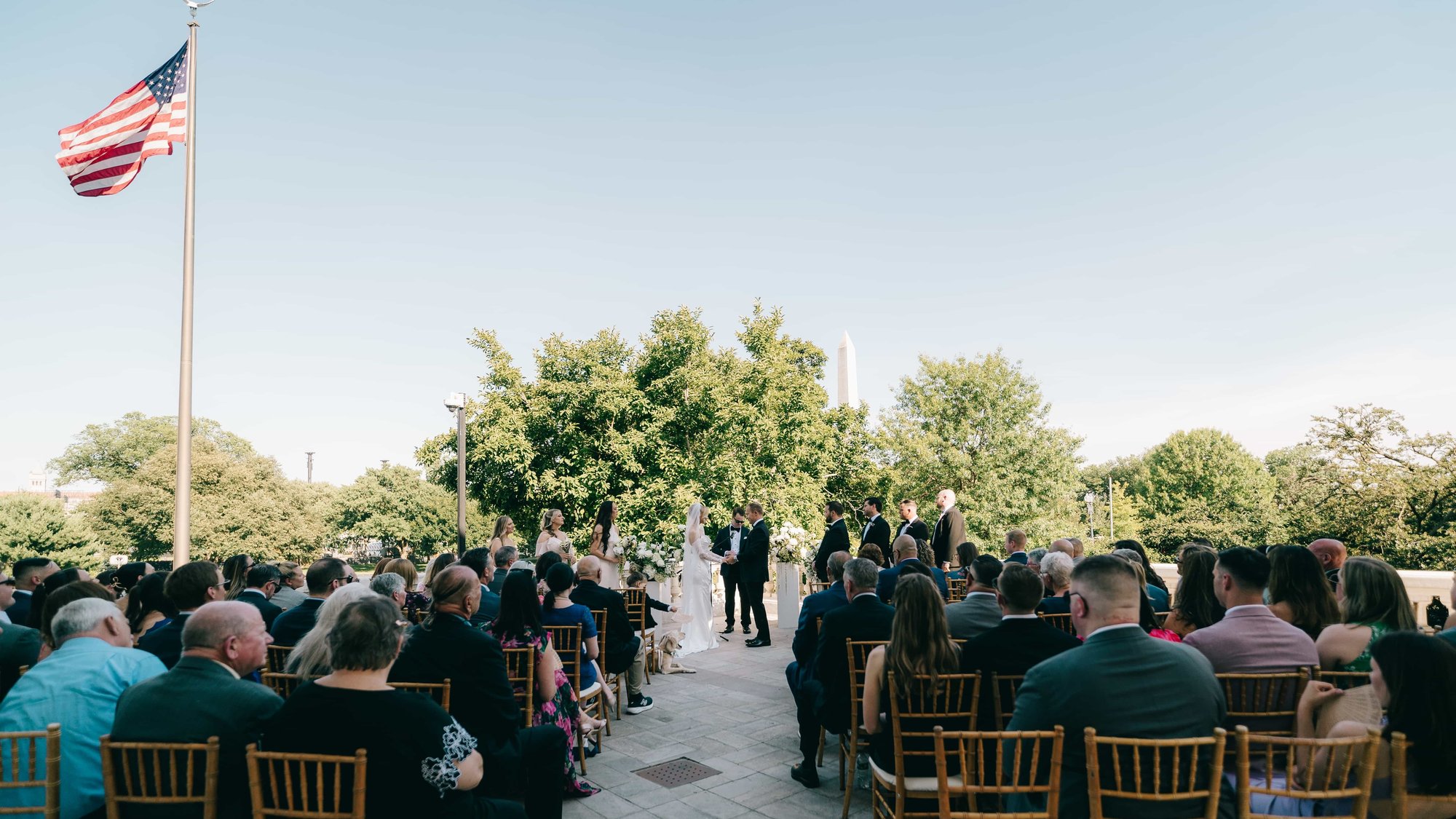 The whole ceremony from the back row, guests seated beneath the flag, the couple far ahead and the Monument over the trees