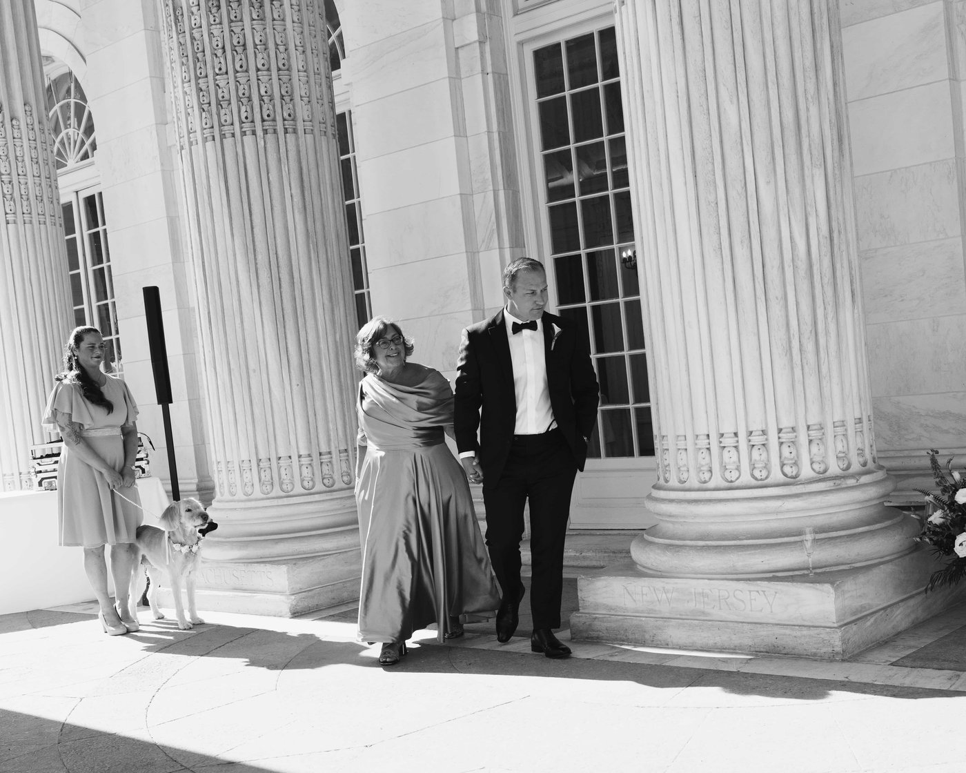 Black and white, a woman in a satin gown walked in on a tuxedoed arm, a dog watching from its lead