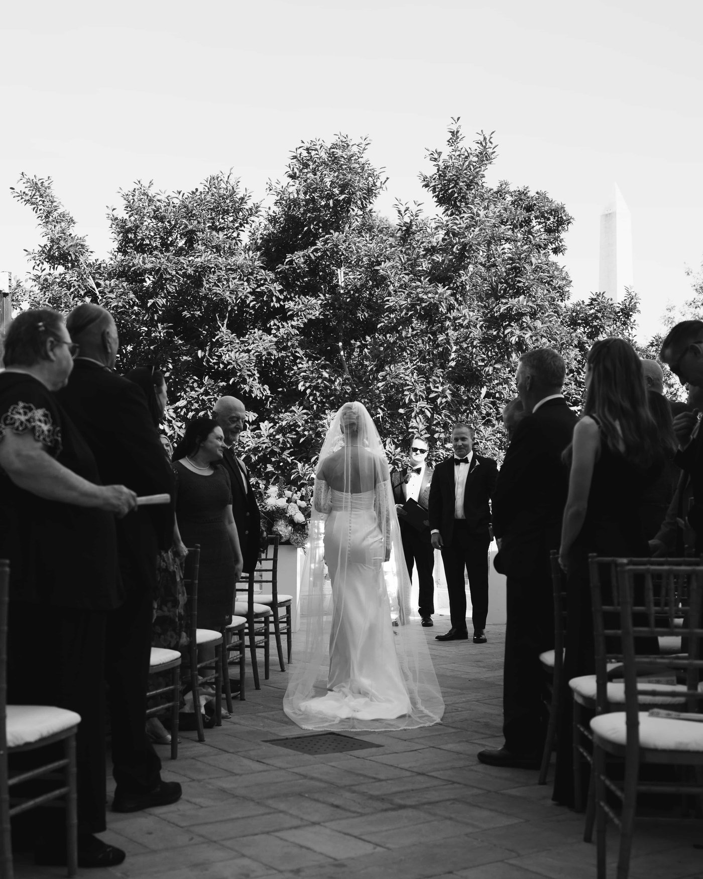 Black and white, the bride coming down the aisle in her long veil, guests on their feet on either side